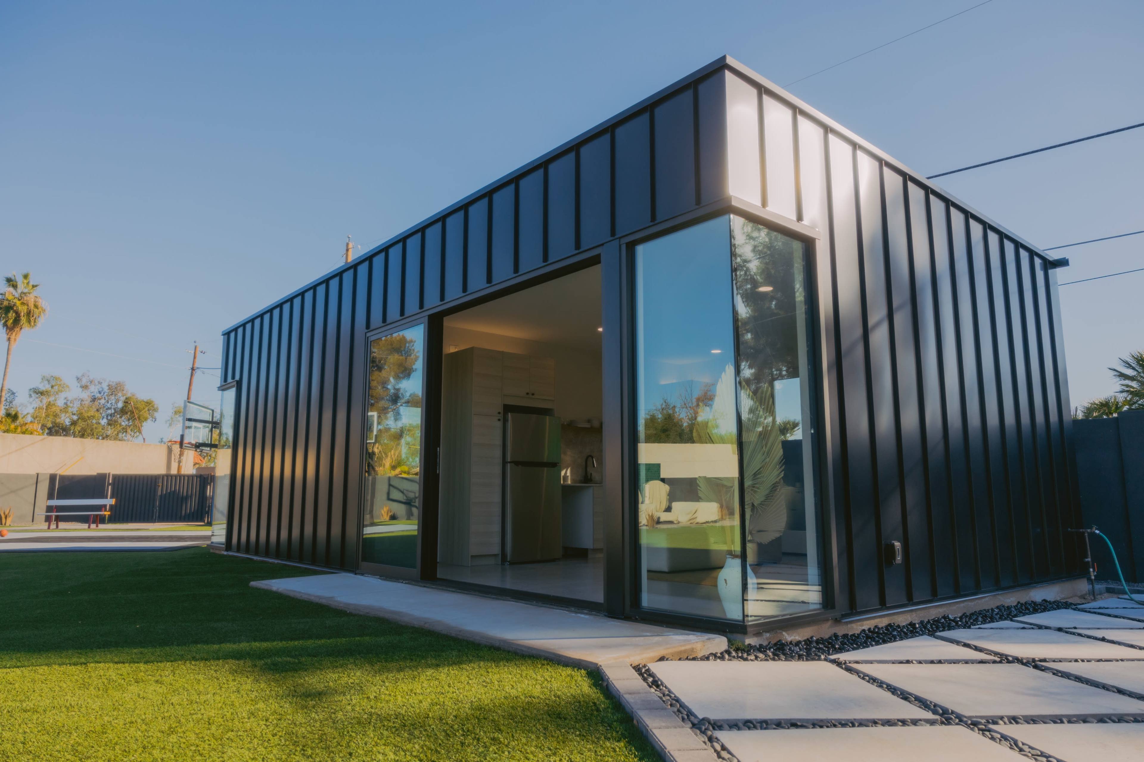 A modern, angular black metal house features large glass windows and is set on a grassy lawn.
