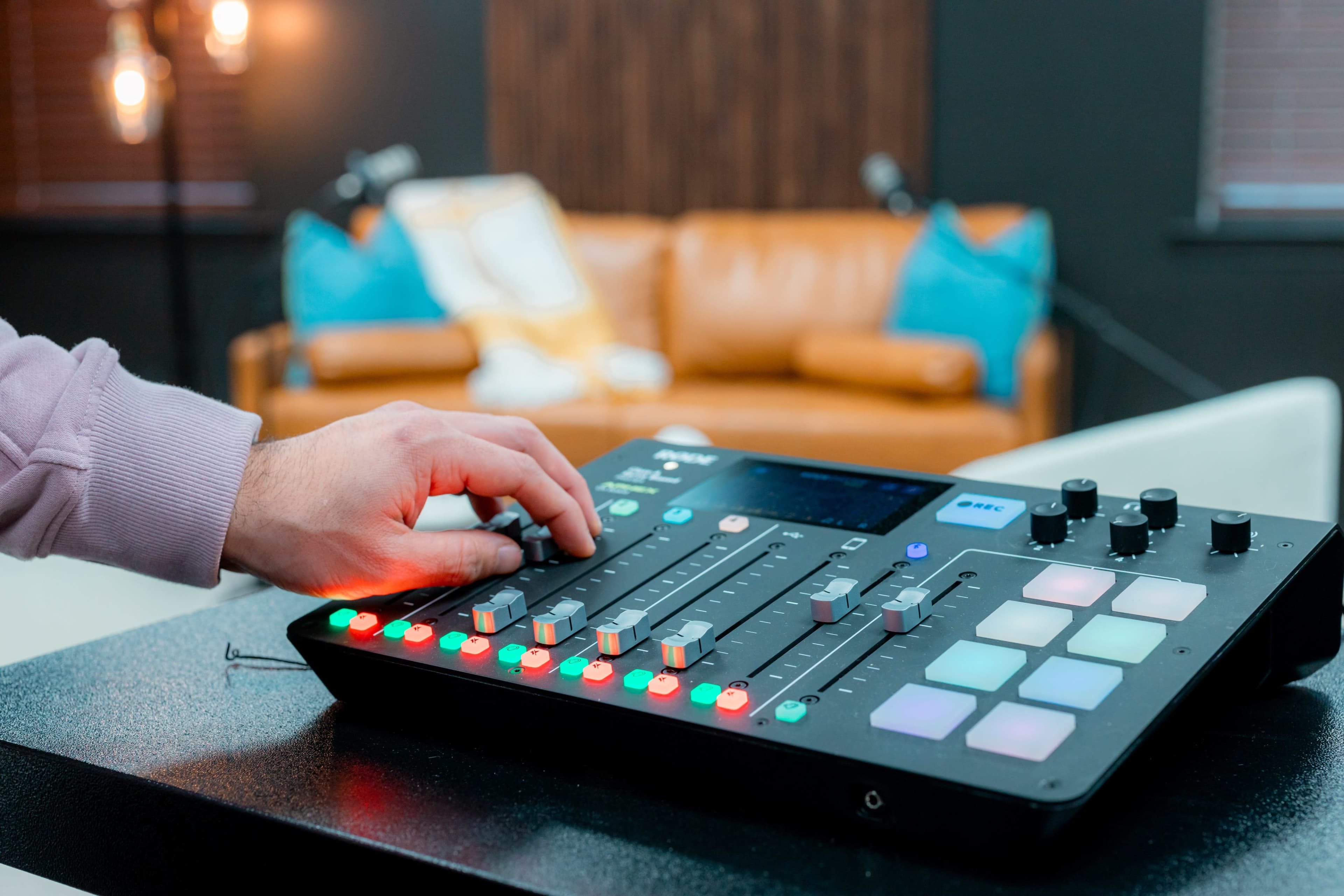 A hand adjusts the controls on a music mixing console in a modern studio setting with a brown leather couch in the background.