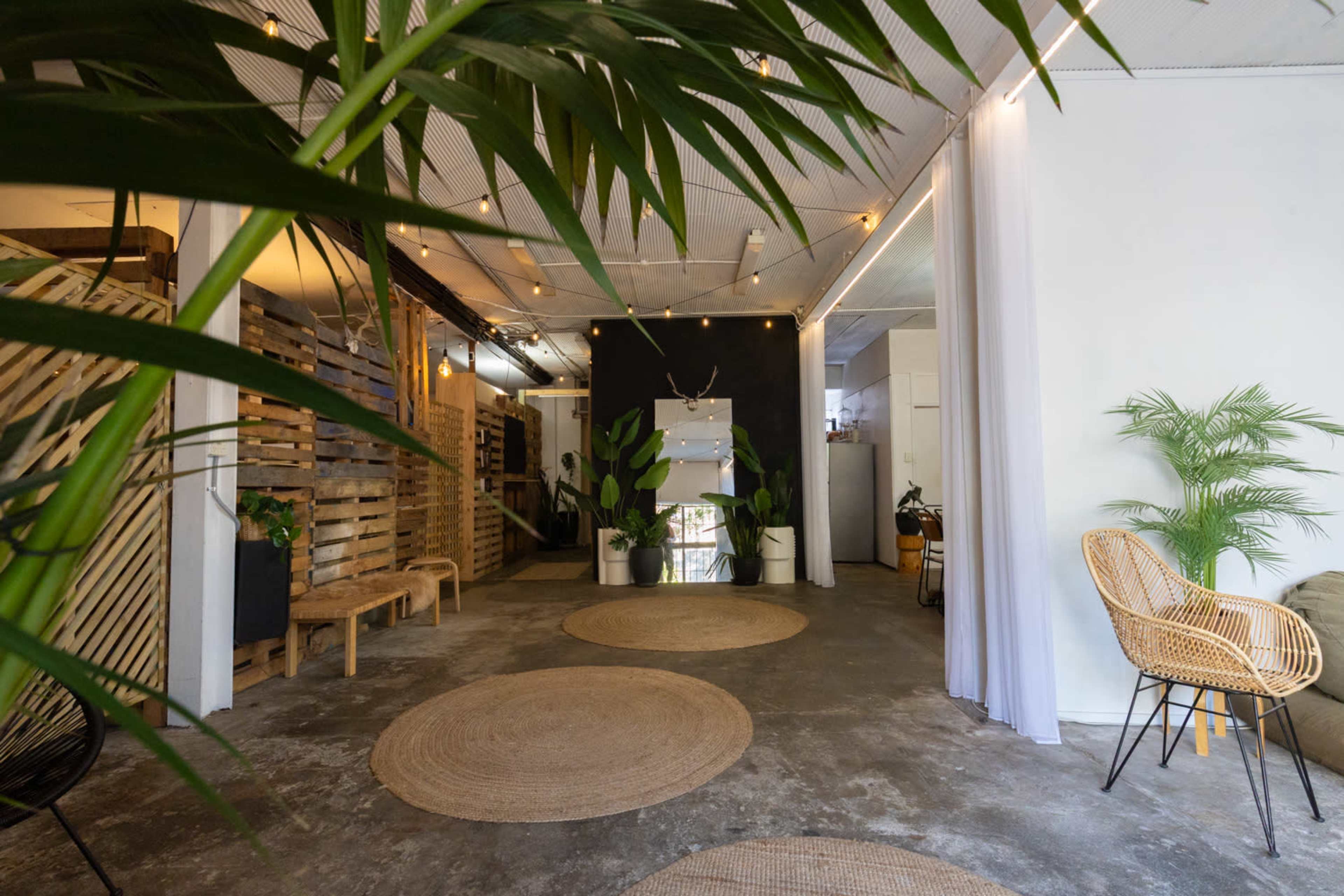 A spacious interior features a combination of wooden and modern decor, with plants, rugs, and seating arrangements in a well-lit area.