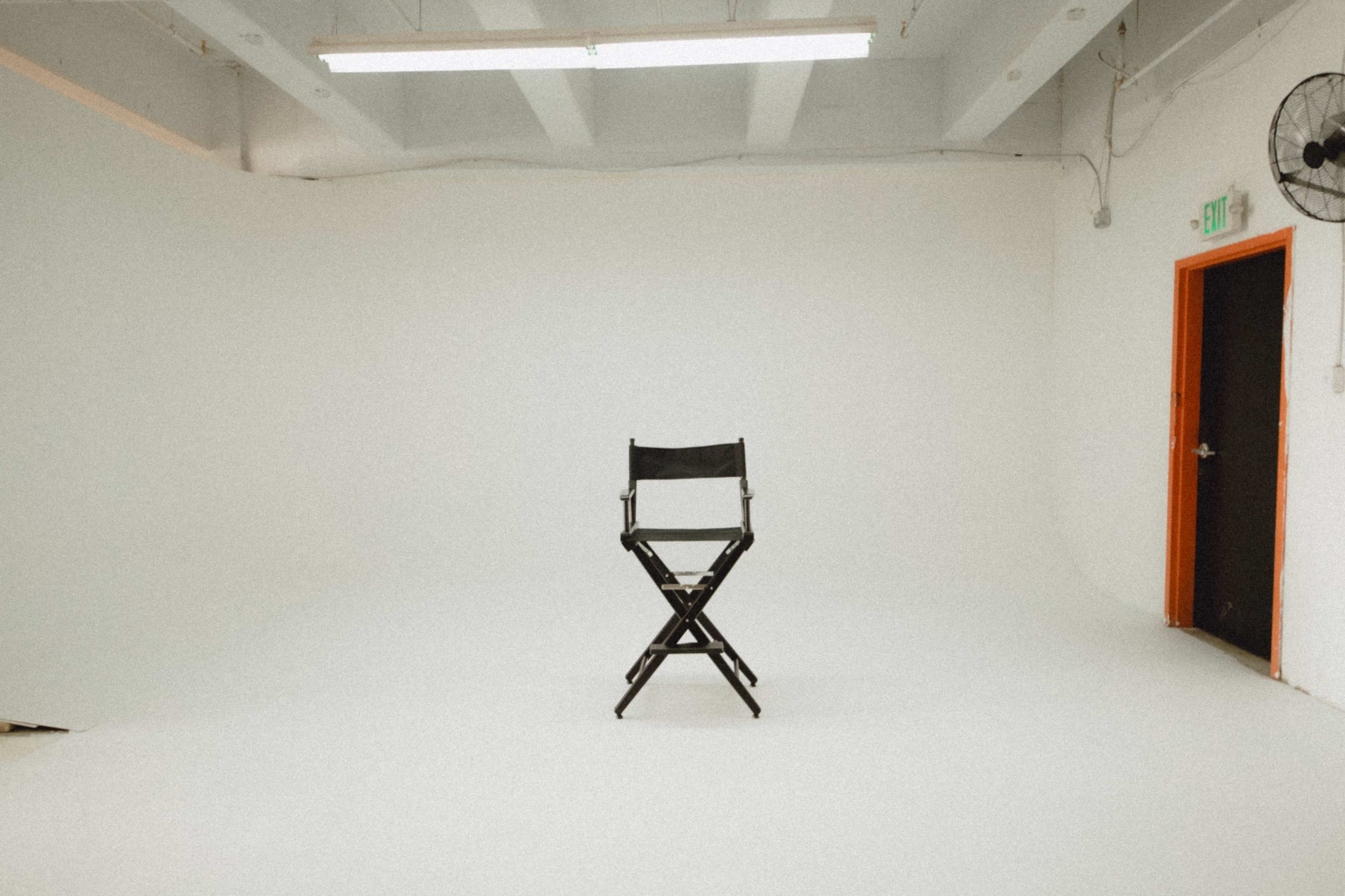 A black director's chair stands alone on a white background in an empty studio.