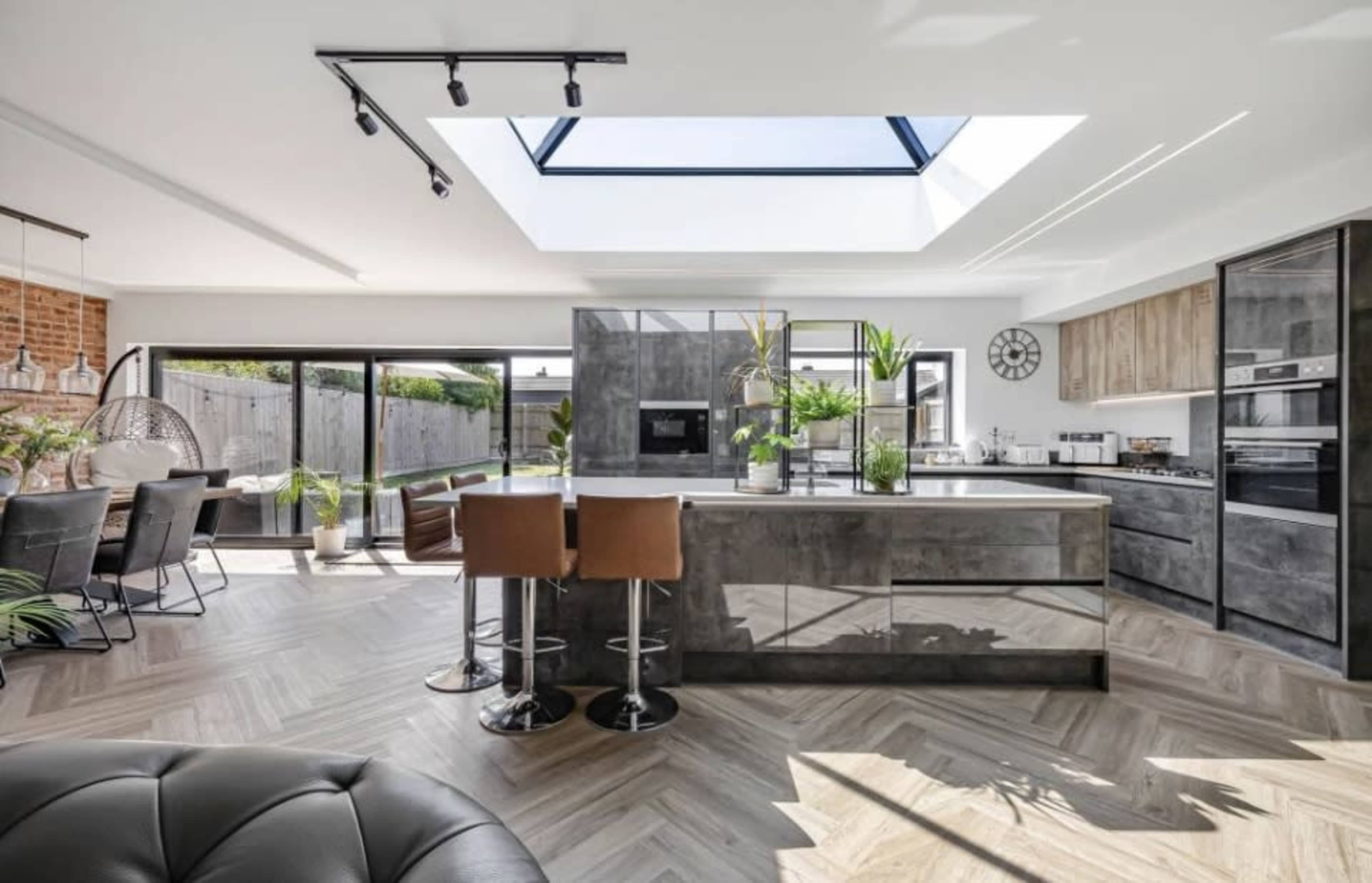 The image shows a modern kitchen with a central island, large windows, and a skylight that allows natural light to illuminate the space.