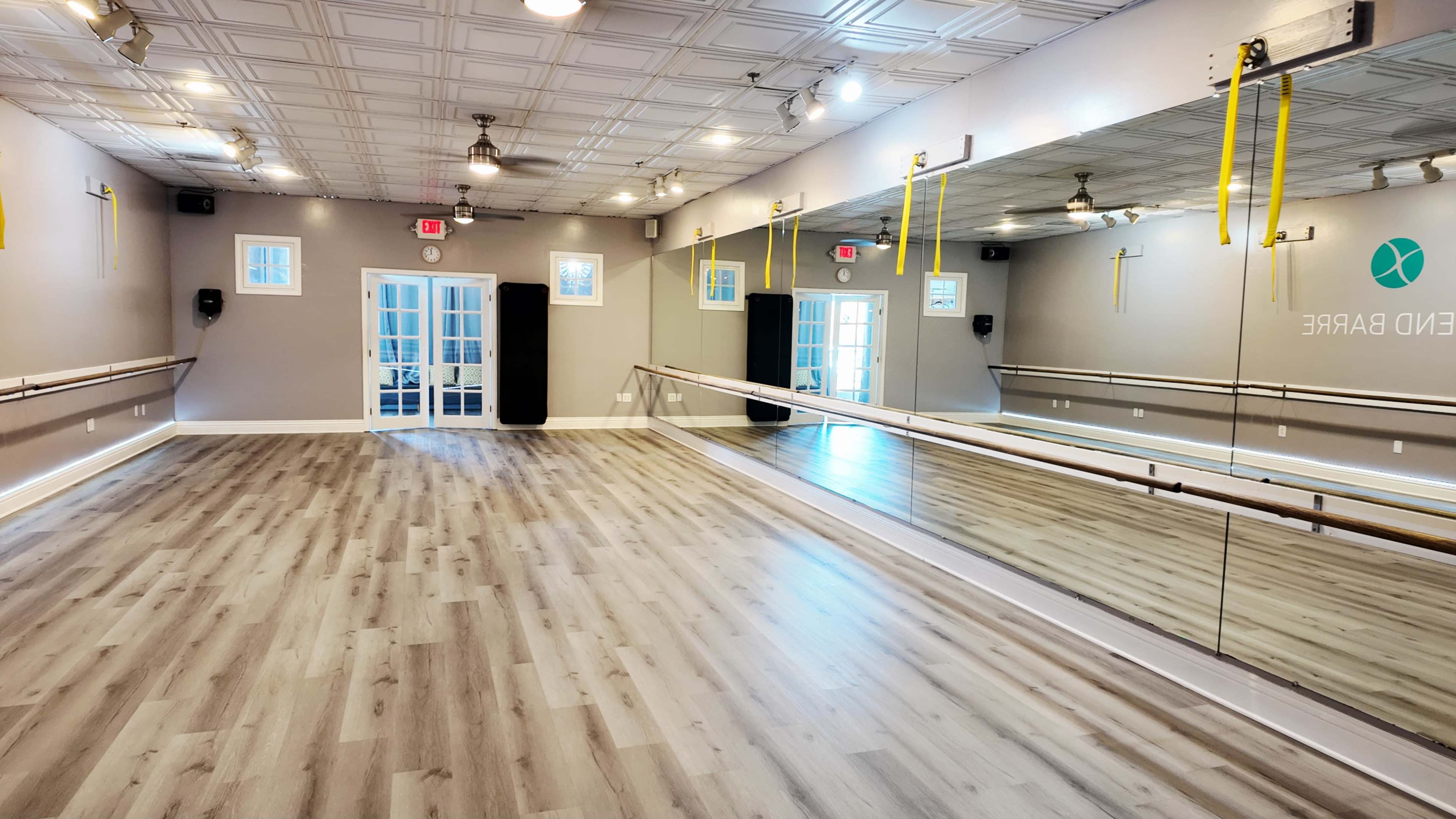The image shows a spacious, well-lit dance studio featuring a large mirror along one wall, ballet barres mounted on the opposite wall, and bright, neutral-colored flooring.