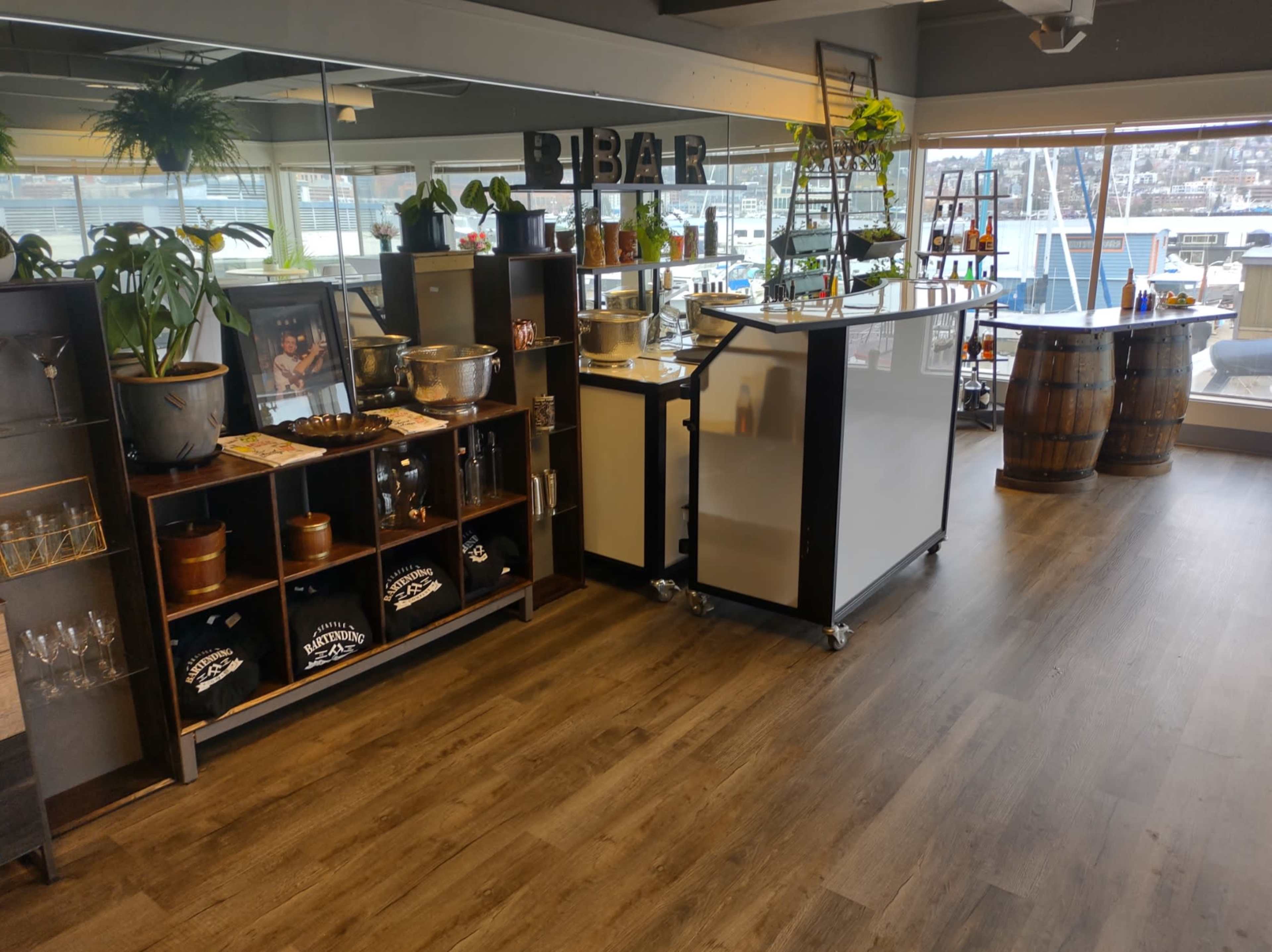 The image shows a well-organized bar area in a modern setting, featuring a counter, shelving with glassware, and decorative plants along with a view of a waterfront.
