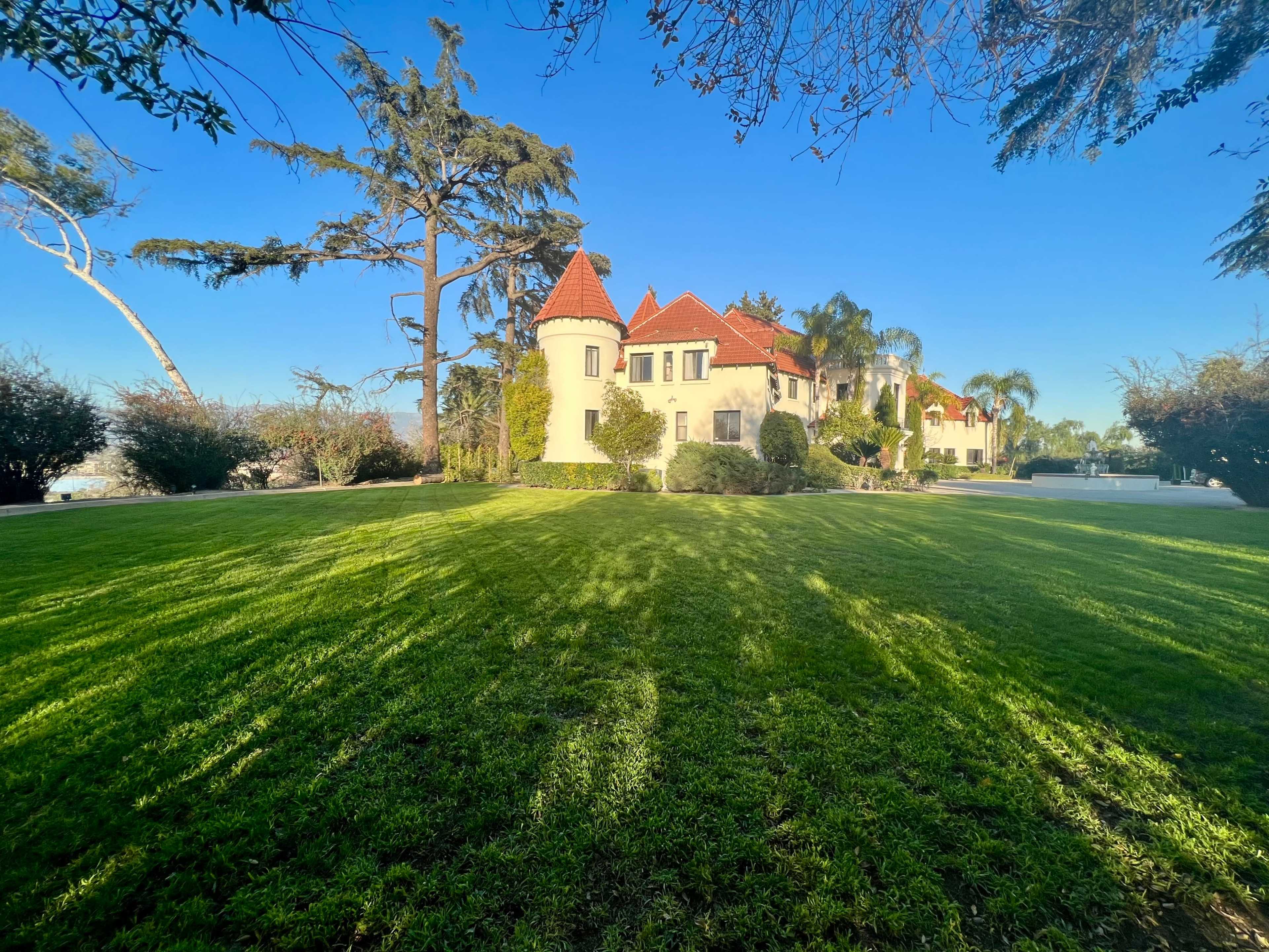 A large, elegant house with red-tiled roofs is surrounded by a well-maintained lawn and trees on a clear day.