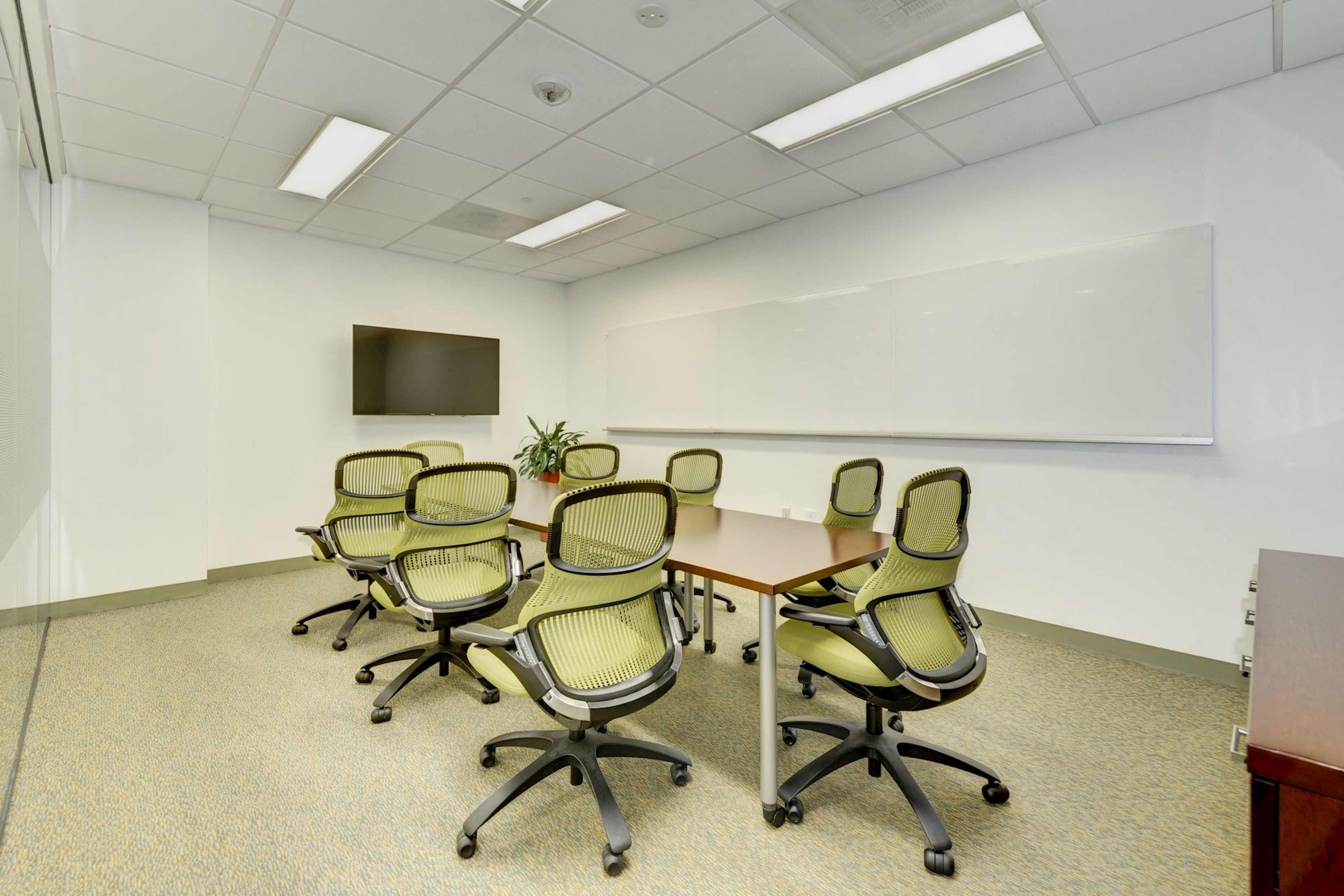 A conference room with a rectangular table, eight rolling chairs, a wall-mounted television, and a dry-erase board.