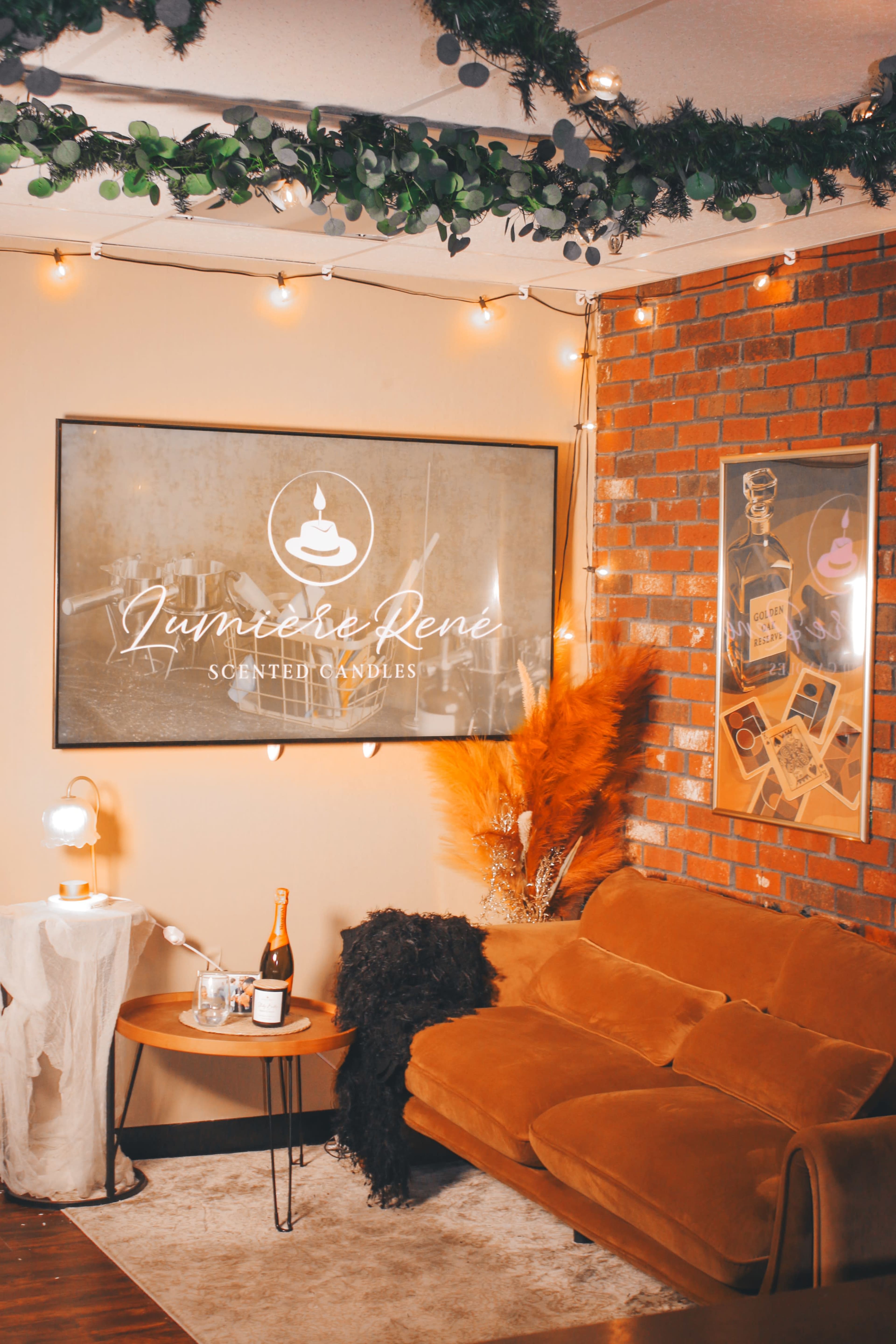 The image features a cozy room with a brown couch, decorative plants, string lights, and a table with a bottle of champagne and a glass.