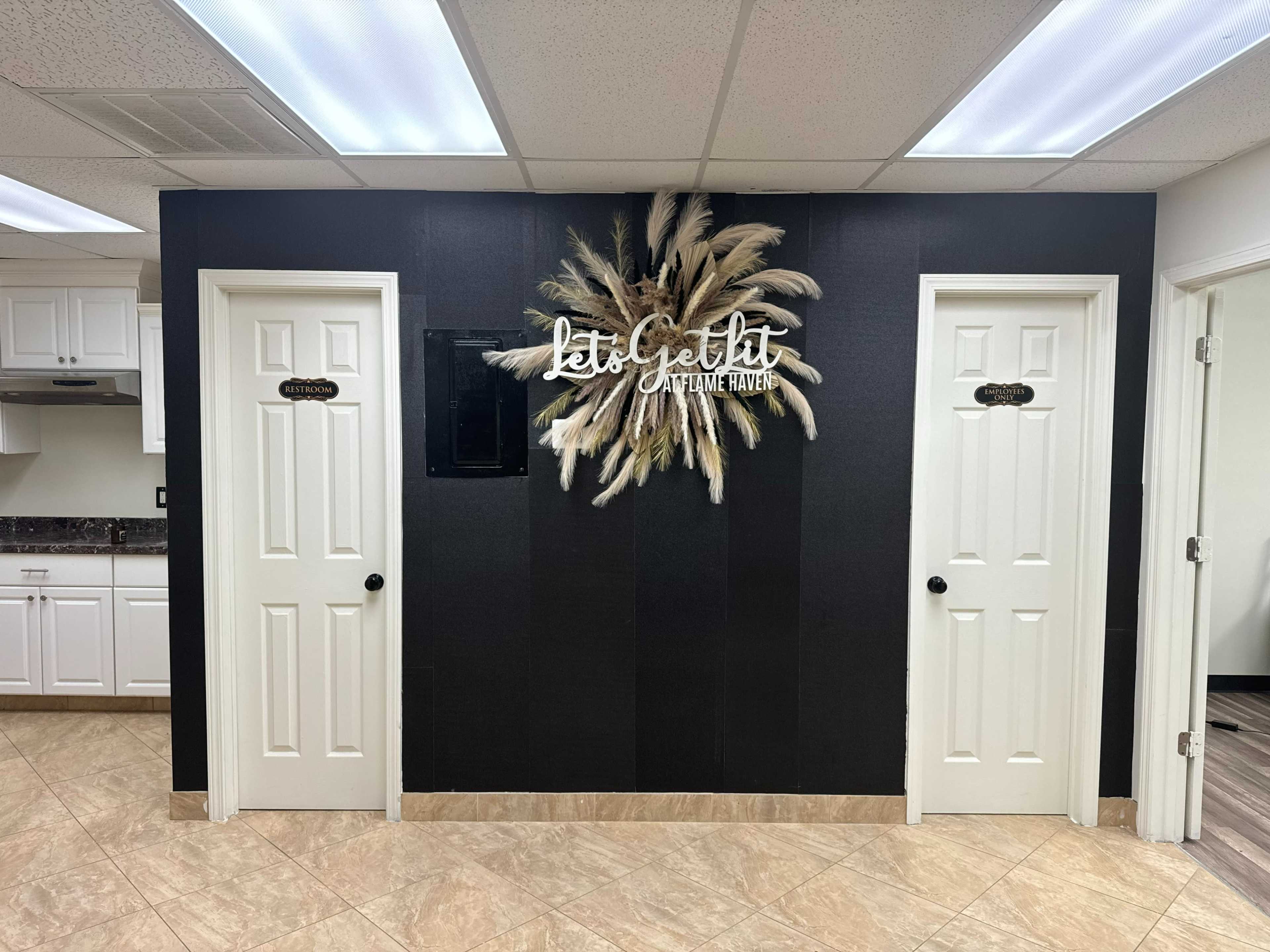 A black wall with a decorative arrangement of pampas grass and a sign, flanked by two white doors, in a room with tile flooring.