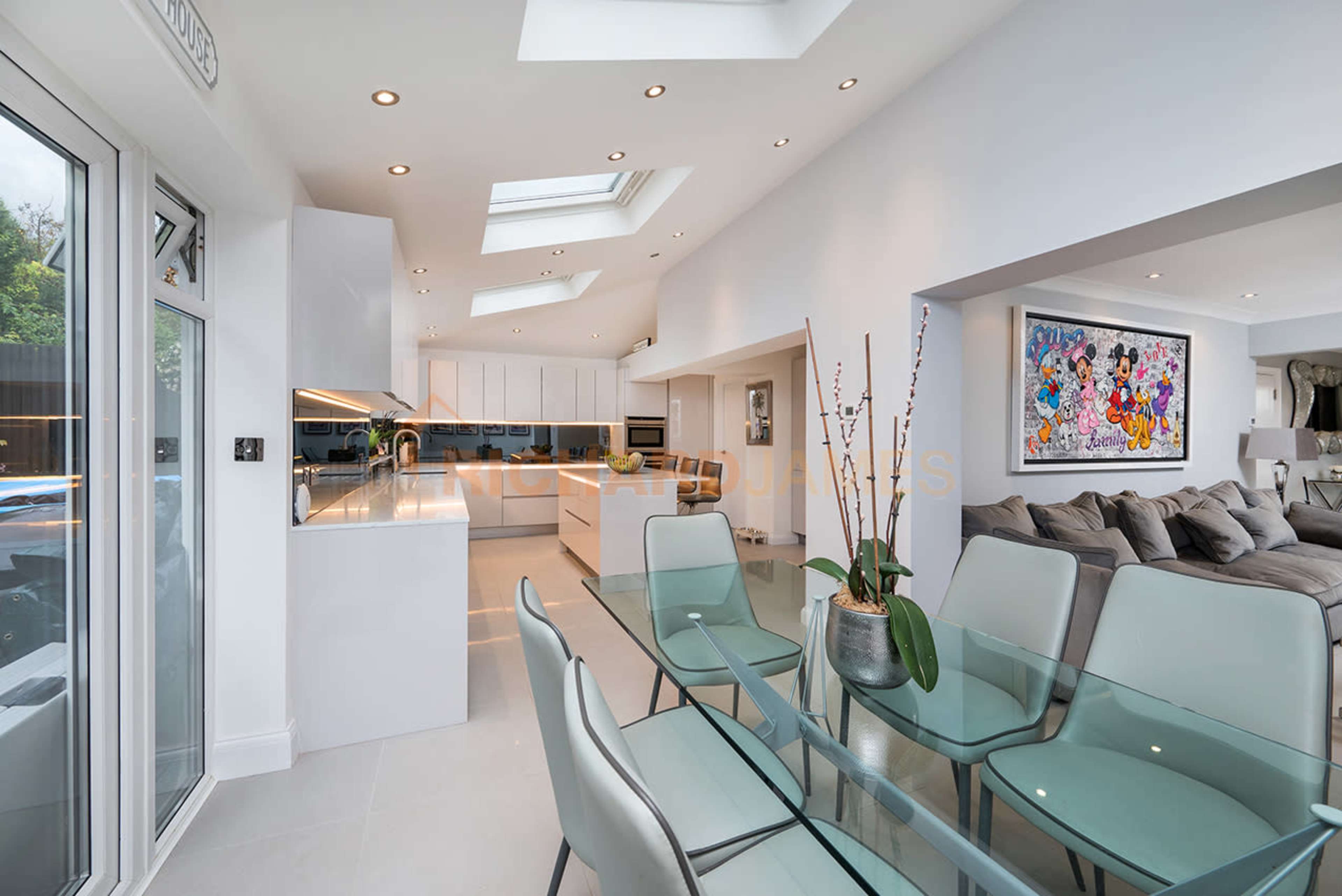 A modern open-plan kitchen and dining area with sleek white cabinetry, large skylights, and a glass dining table surrounded by chairs.