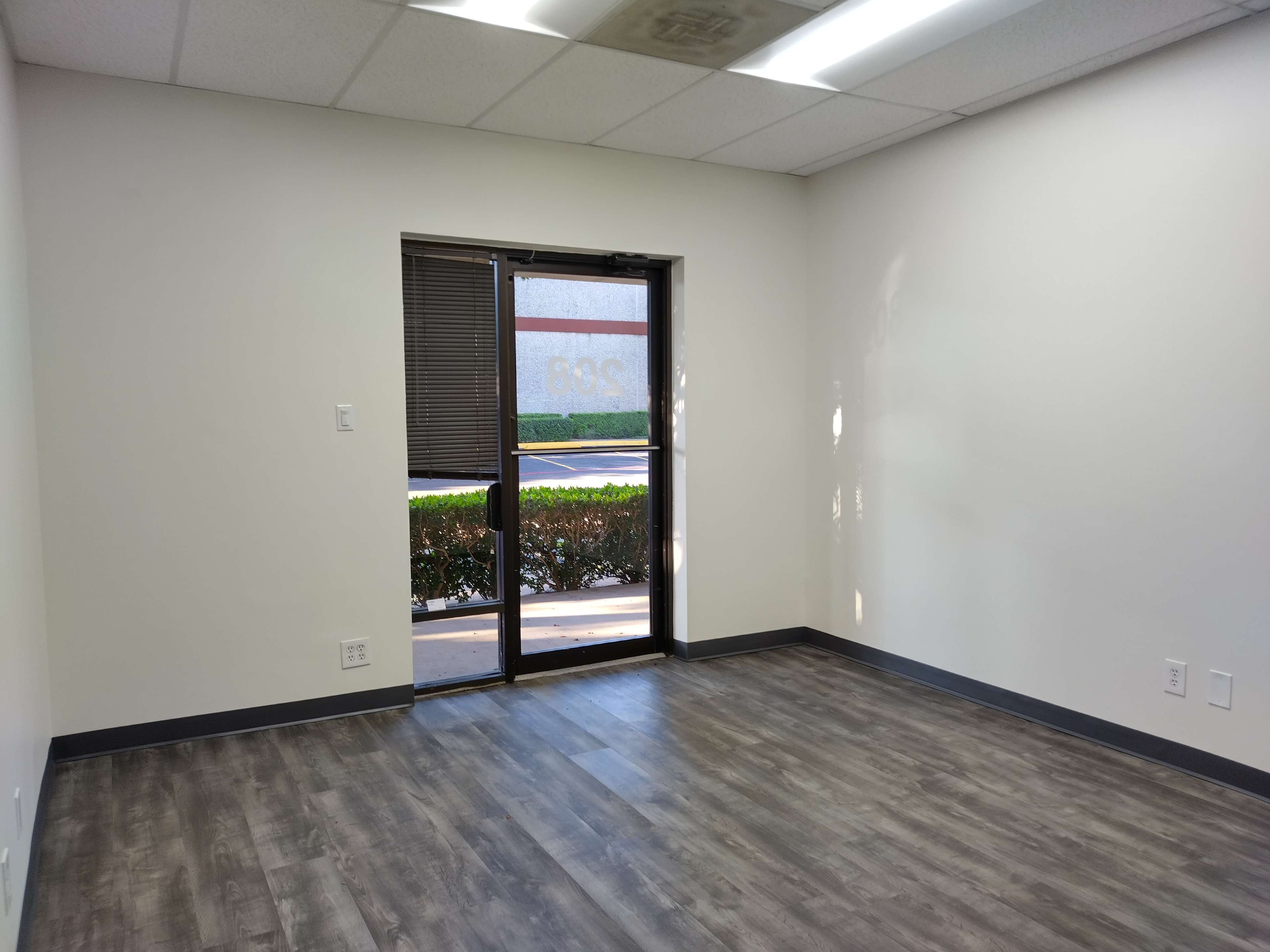 The image shows an empty room with a door leading to an outdoor area, featuring gray flooring and a window with blinds.