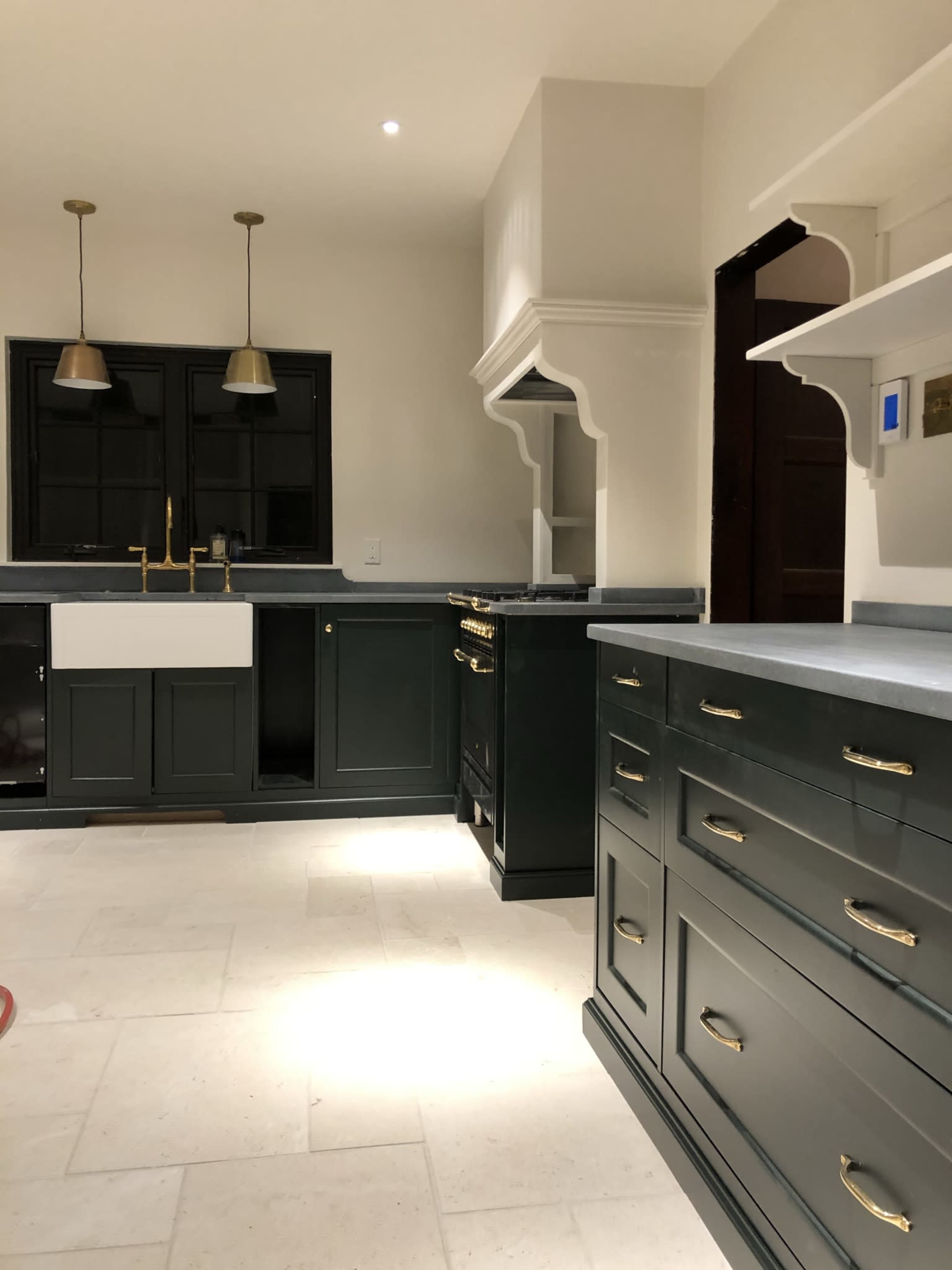 The image shows a modern kitchen featuring dark green cabinets, a central island with drawers, a farmhouse sink, and gold pendant lights.