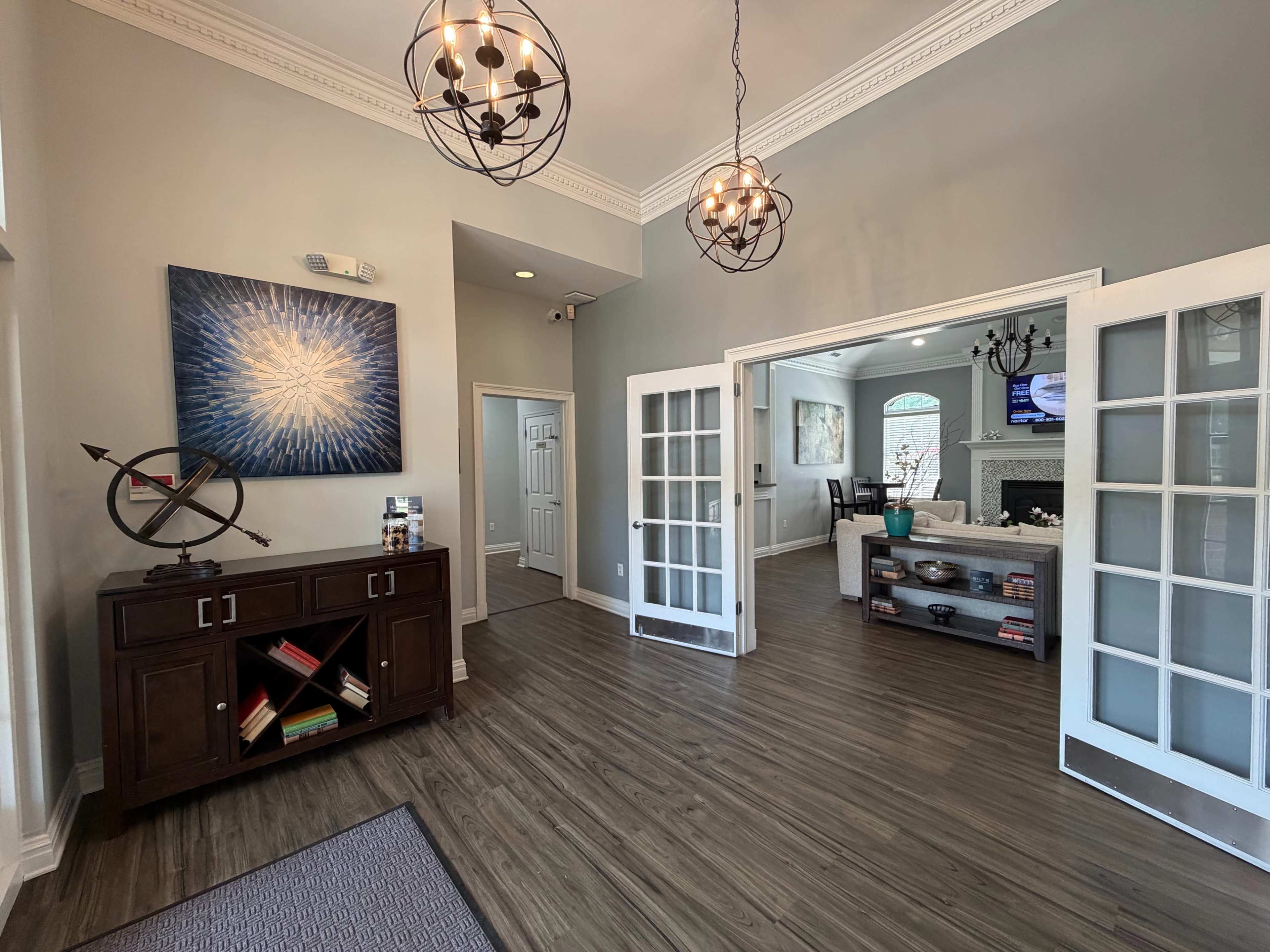 The image shows a well-lit entryway with a wooden console table, a large abstract painting on the wall, and French doors leading to a living area.