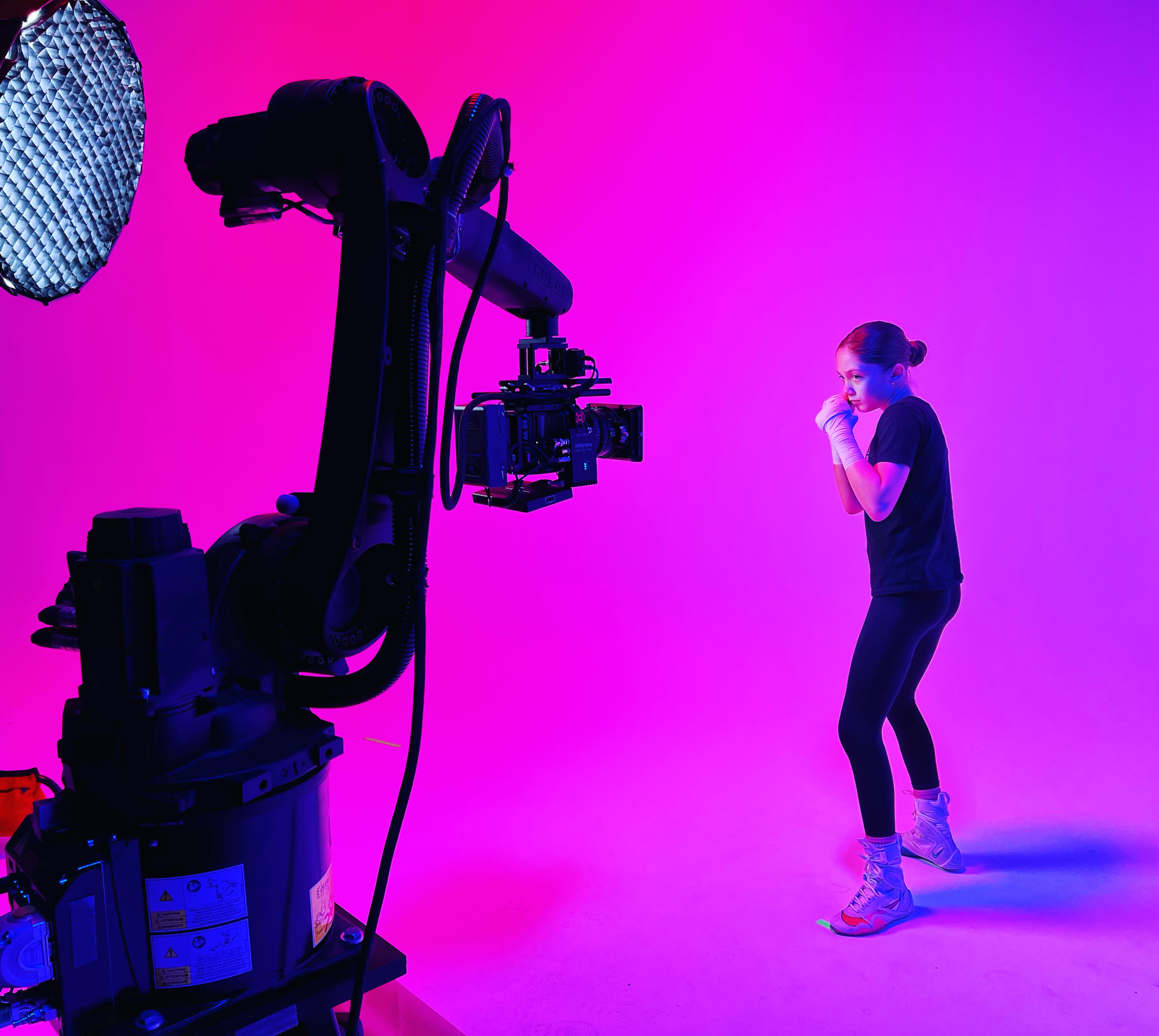 A person in athletic clothing is positioned in a boxing stance on a brightly lit stage with a robotic camera arm capturing the scene.