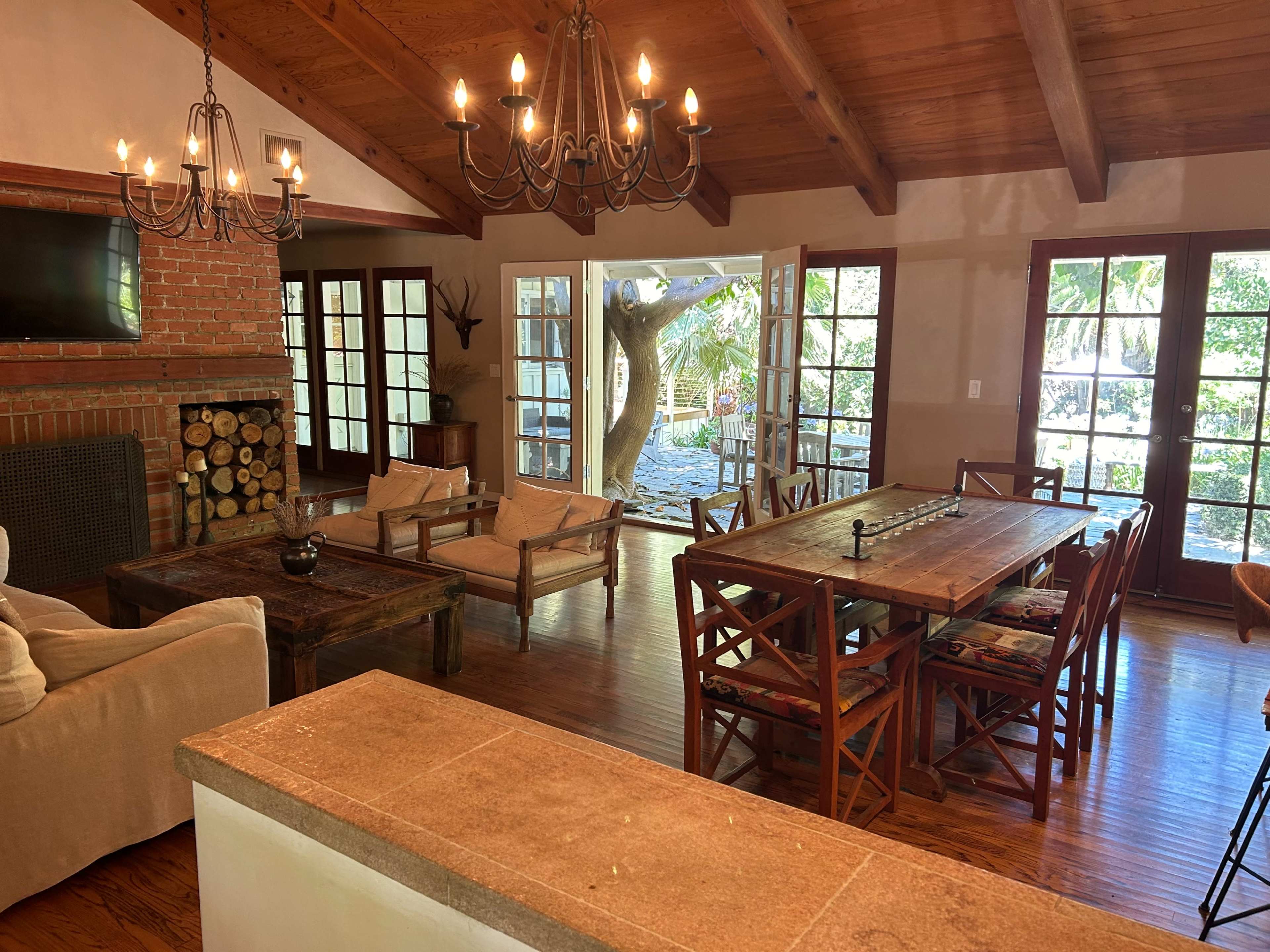 A spacious living area with a wooden ceiling, a large dining table, comfortable seating, and windows that open to an outdoor space with trees.