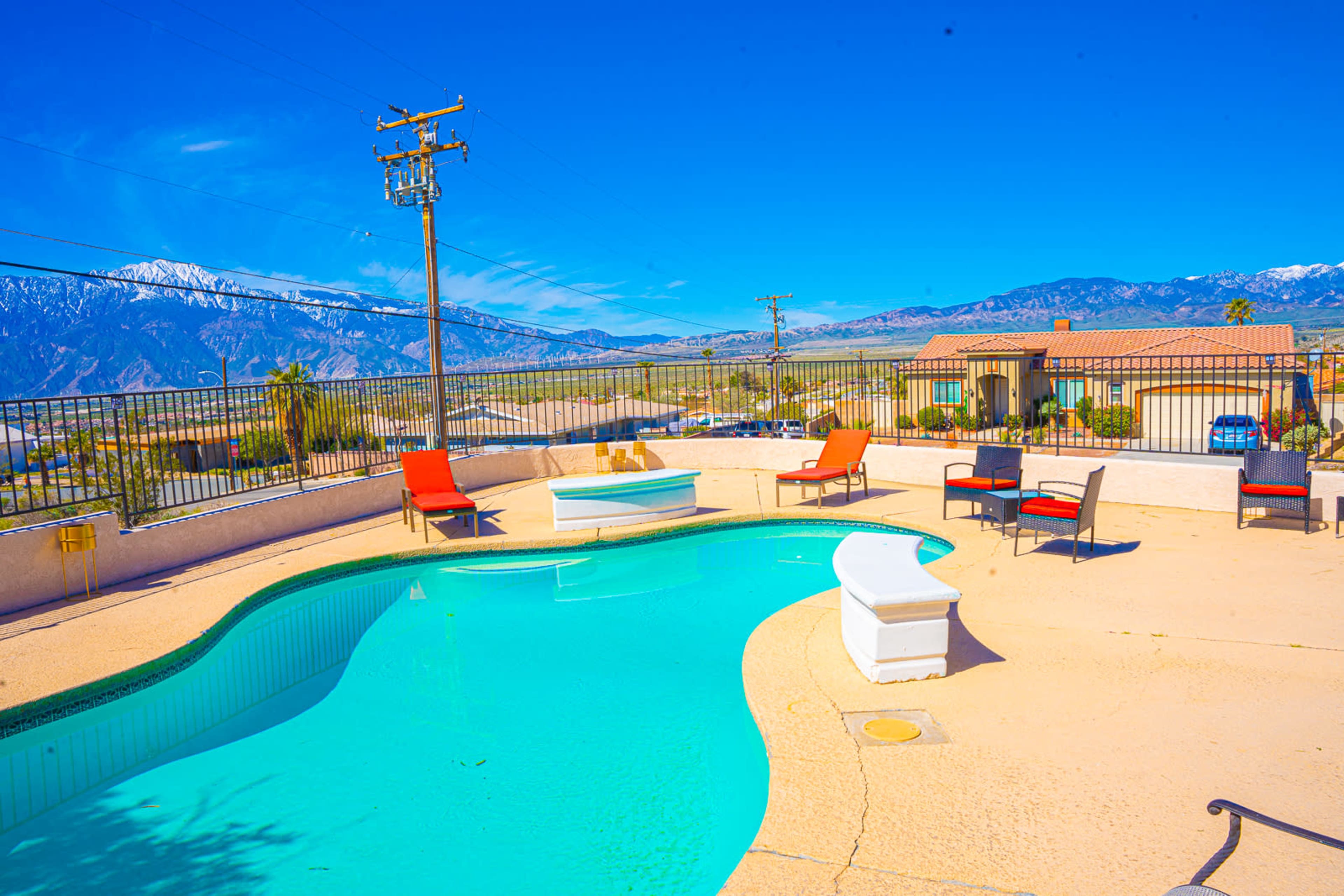 A swimming pool with a mountainous backdrop is surrounded by chairs and lounge areas under a clear blue sky.