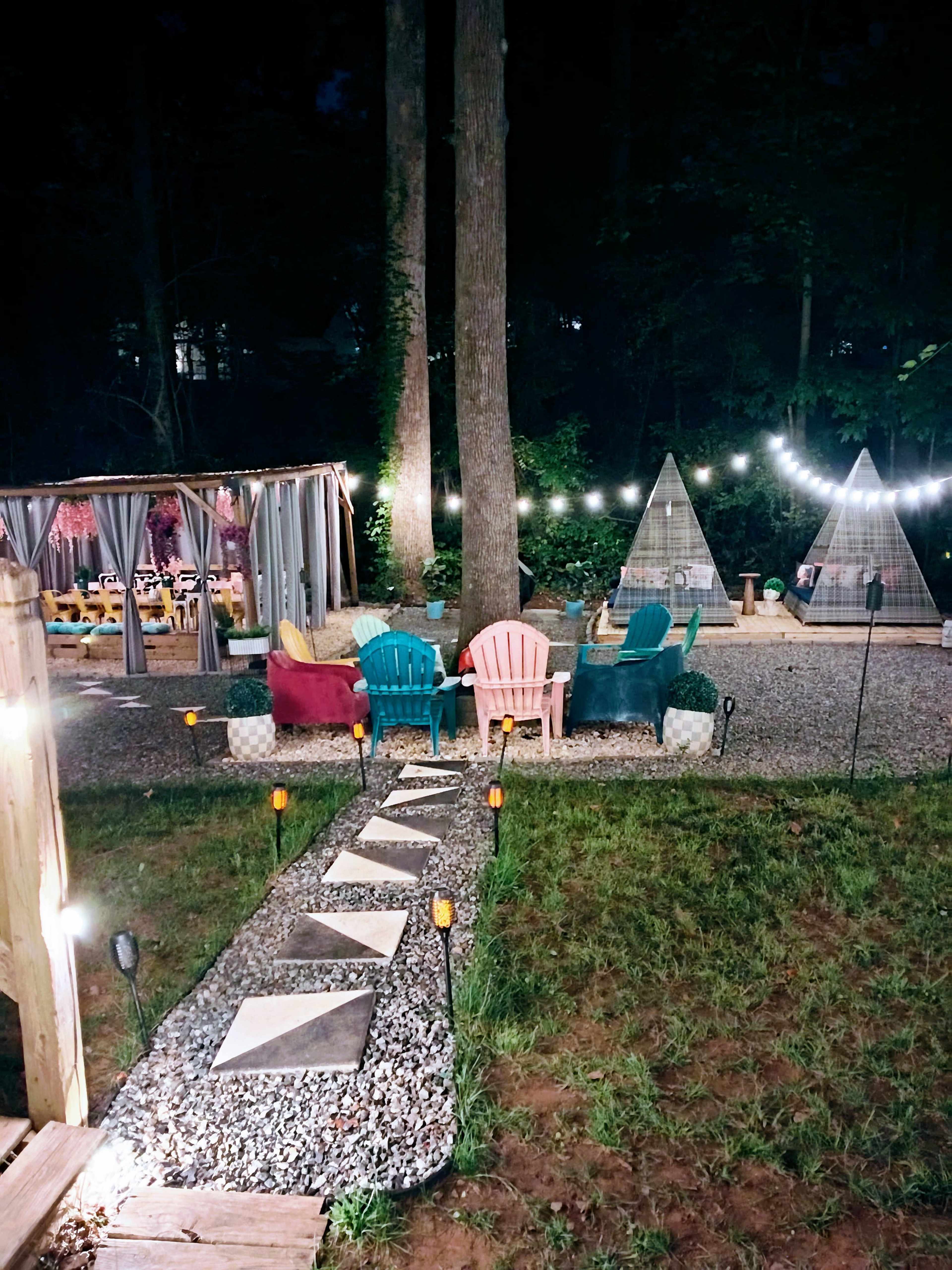 The scene features a backyard at night with colorful chairs, stone pathways, and illuminated decorations leading to two pyramid-shaped tents.
