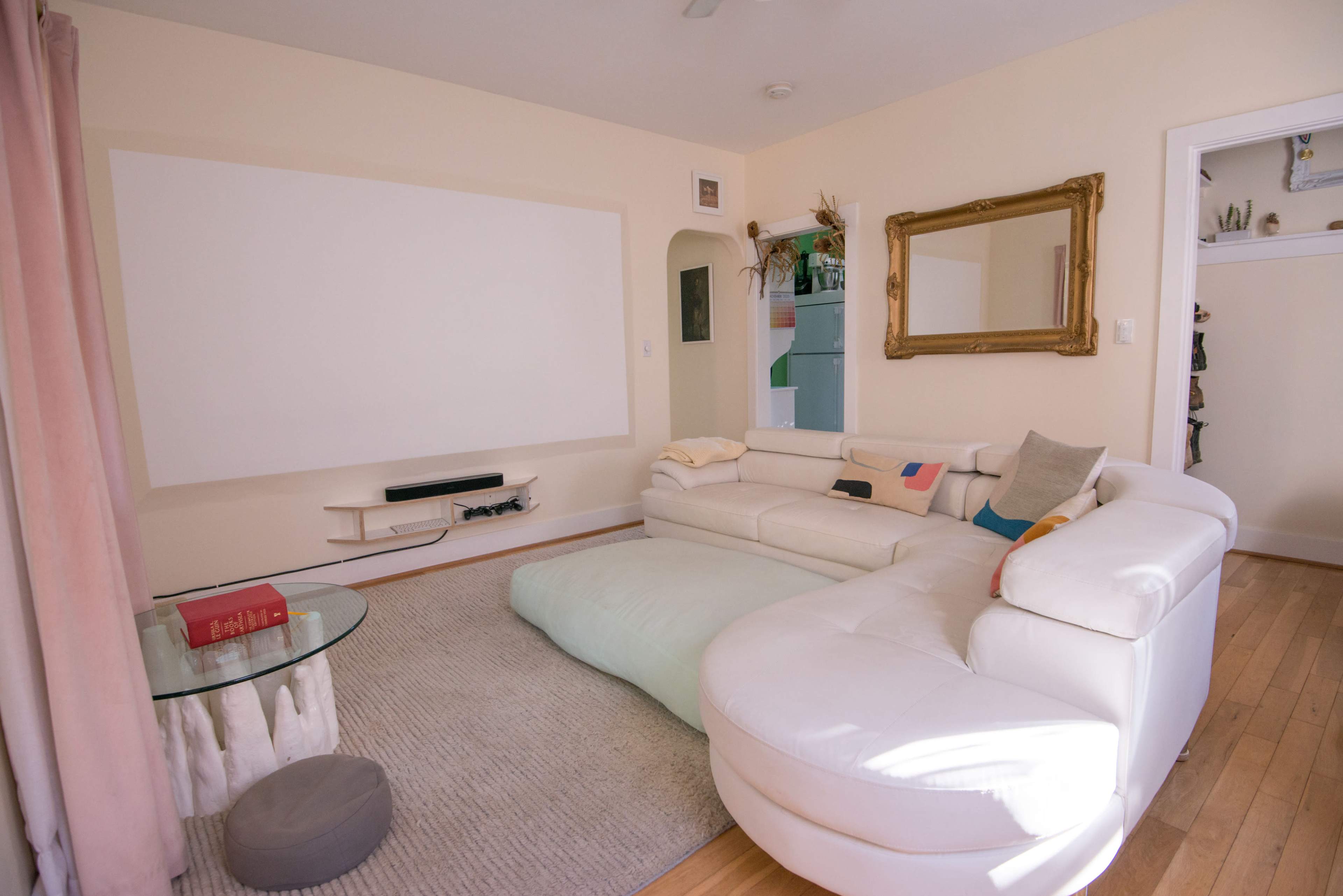 The image shows a living room with a large white sectional sofa, a glass coffee table, a wall-mounted flat-screen TV, and a blank wall intended for a projector screen.