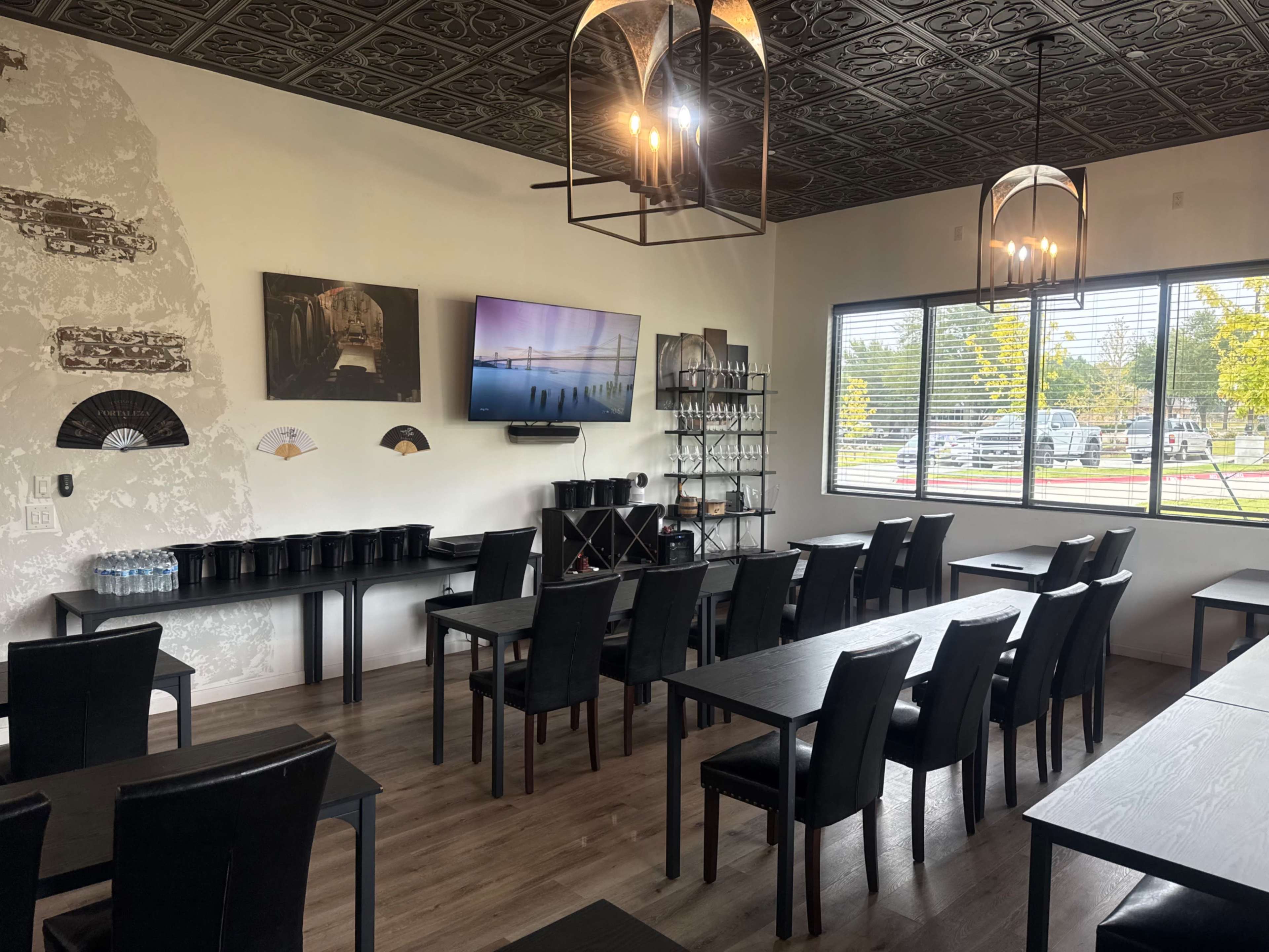 The image shows a neat meeting room furnished with several black tables and chairs, a television mounted on the wall, and large windows allowing natural light to enter.