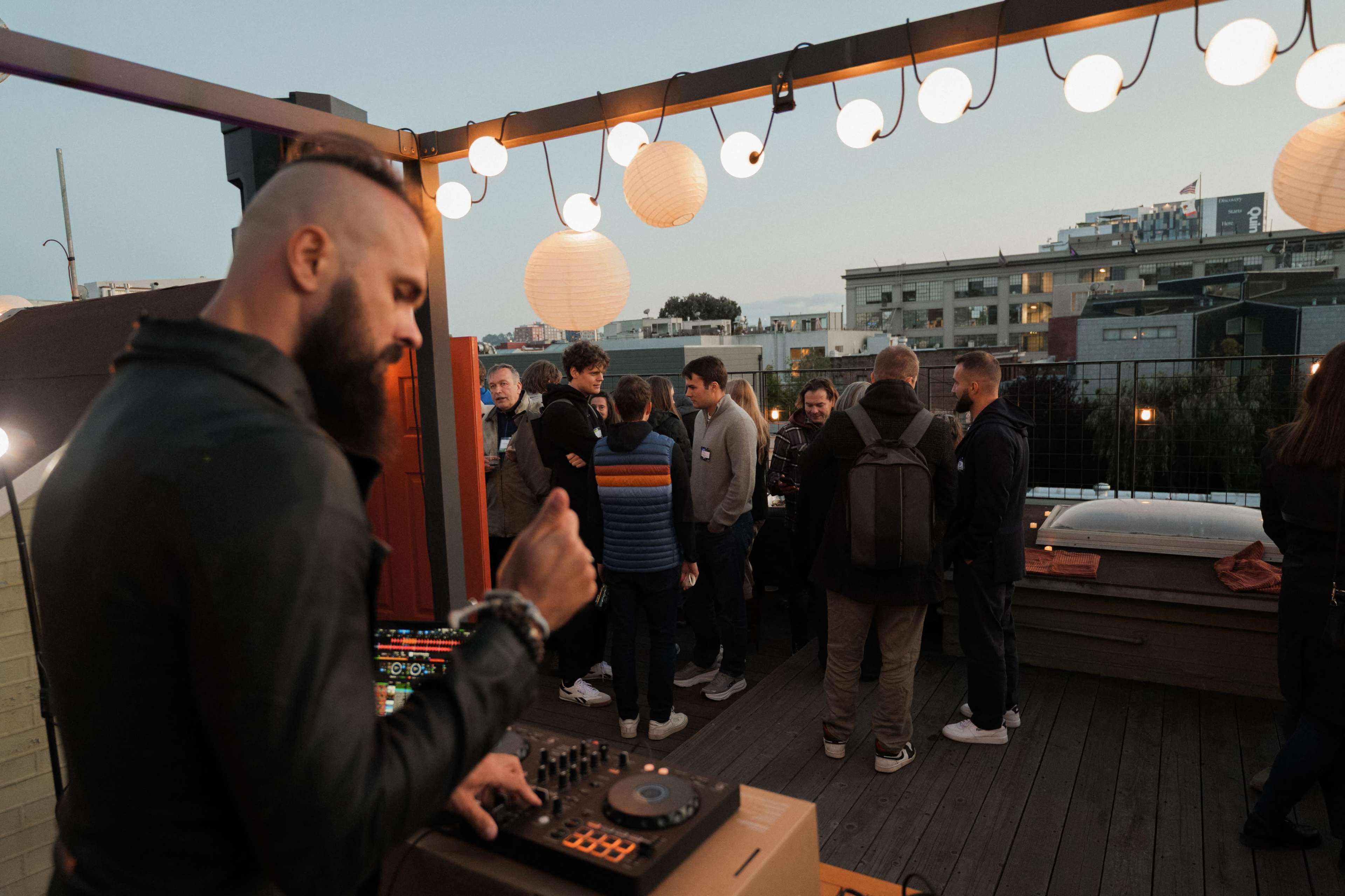 A man with a beard operates a DJ setup on a rooftop as a group of people socialize in the background under lantern lights.
