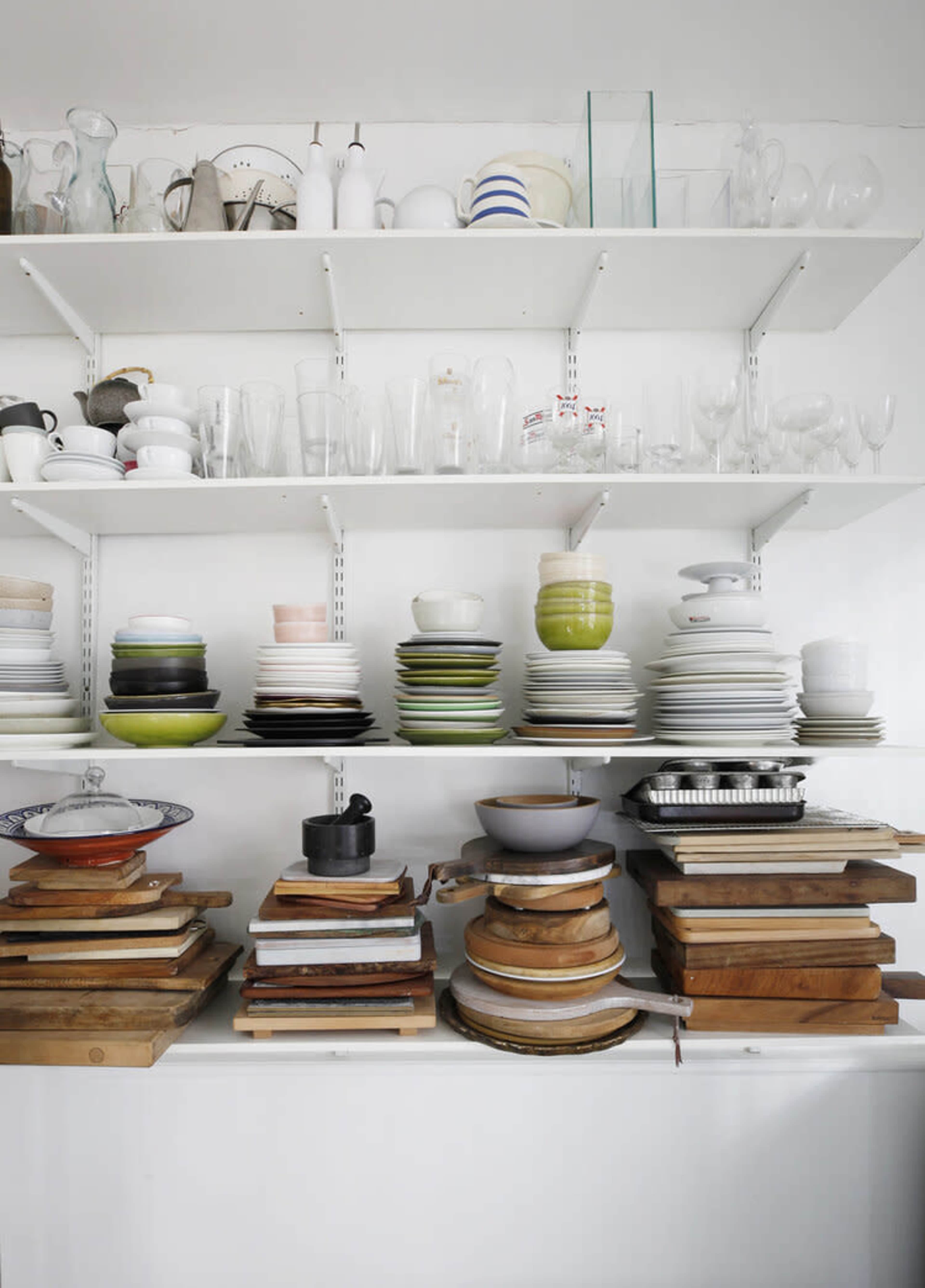 A variety of dishes, glassware, and cutting boards are neatly arranged on white shelves in a minimalist kitchen setting.