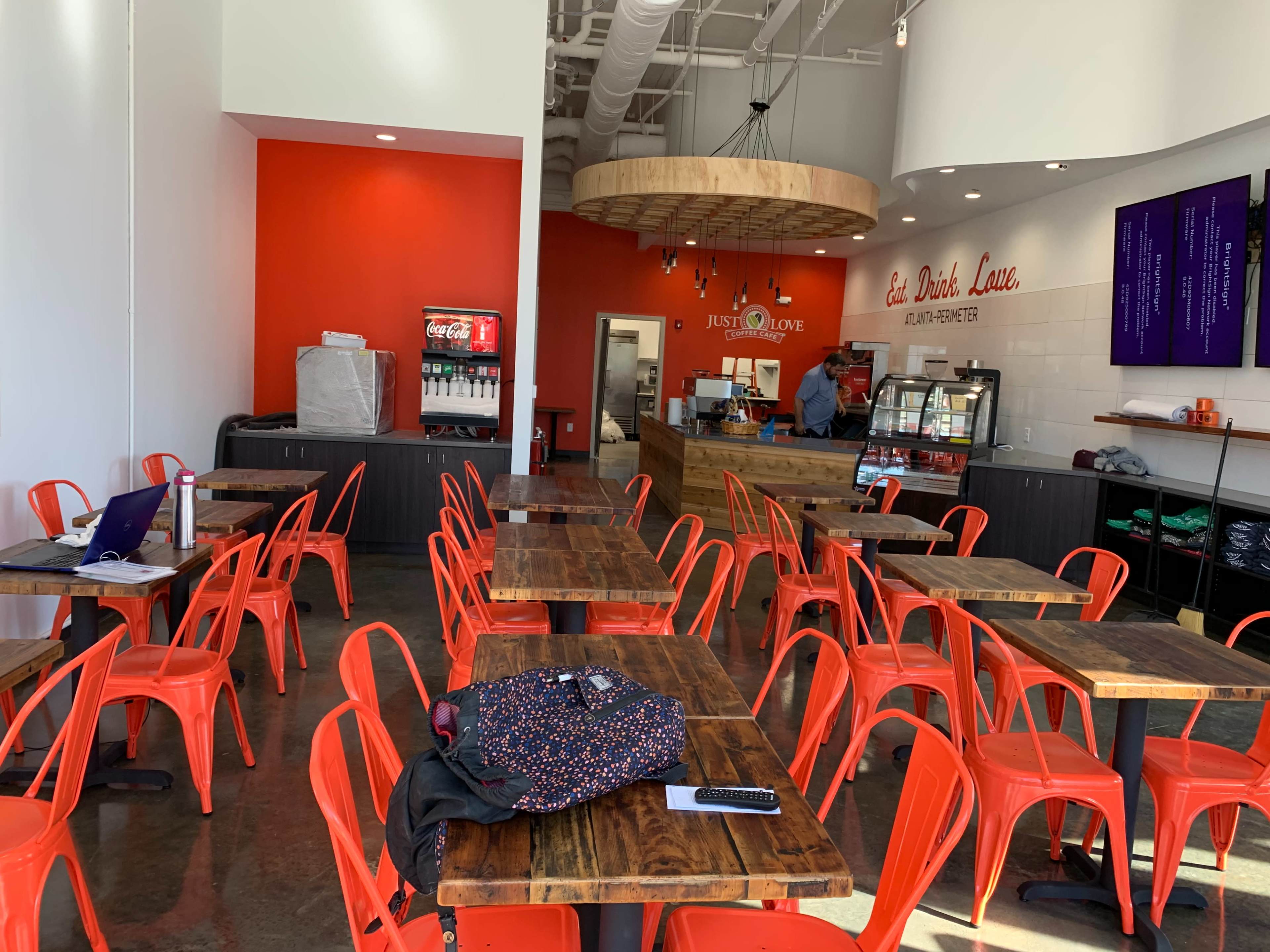 The image shows a casual dining space with wooden tables and red metal chairs, featuring a service counter and a drink station in a brightly lit setting.