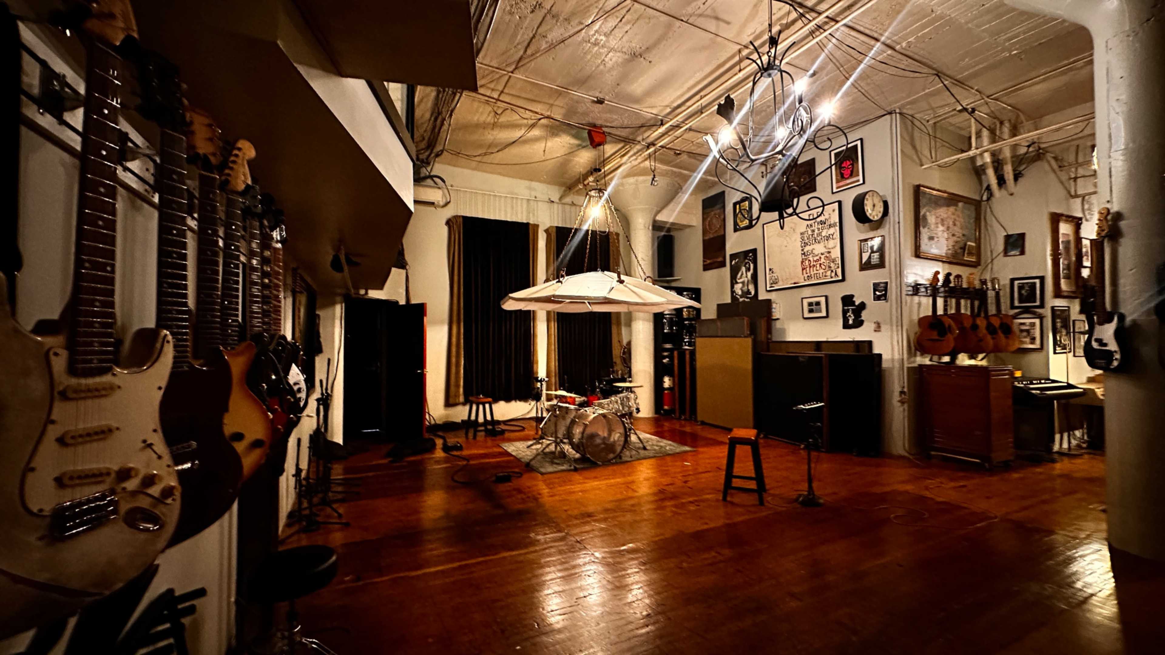 The image shows a music studio featuring guitars on the walls, a drum set in the center, and various instruments and vintage decor around the room.