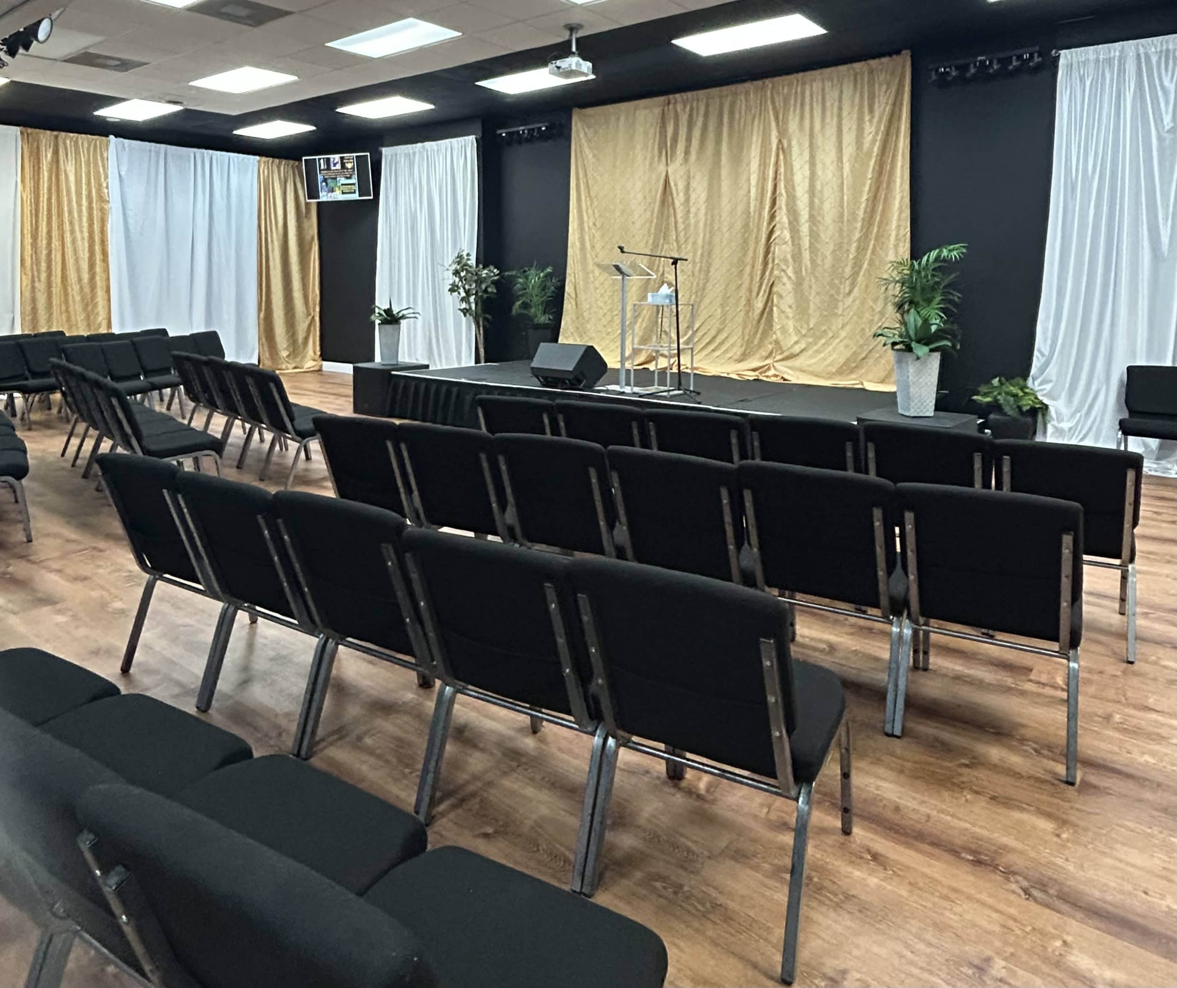The image shows an auditorium-style room with black chairs arranged in rows facing a small stage decorated with white and gold drapes.