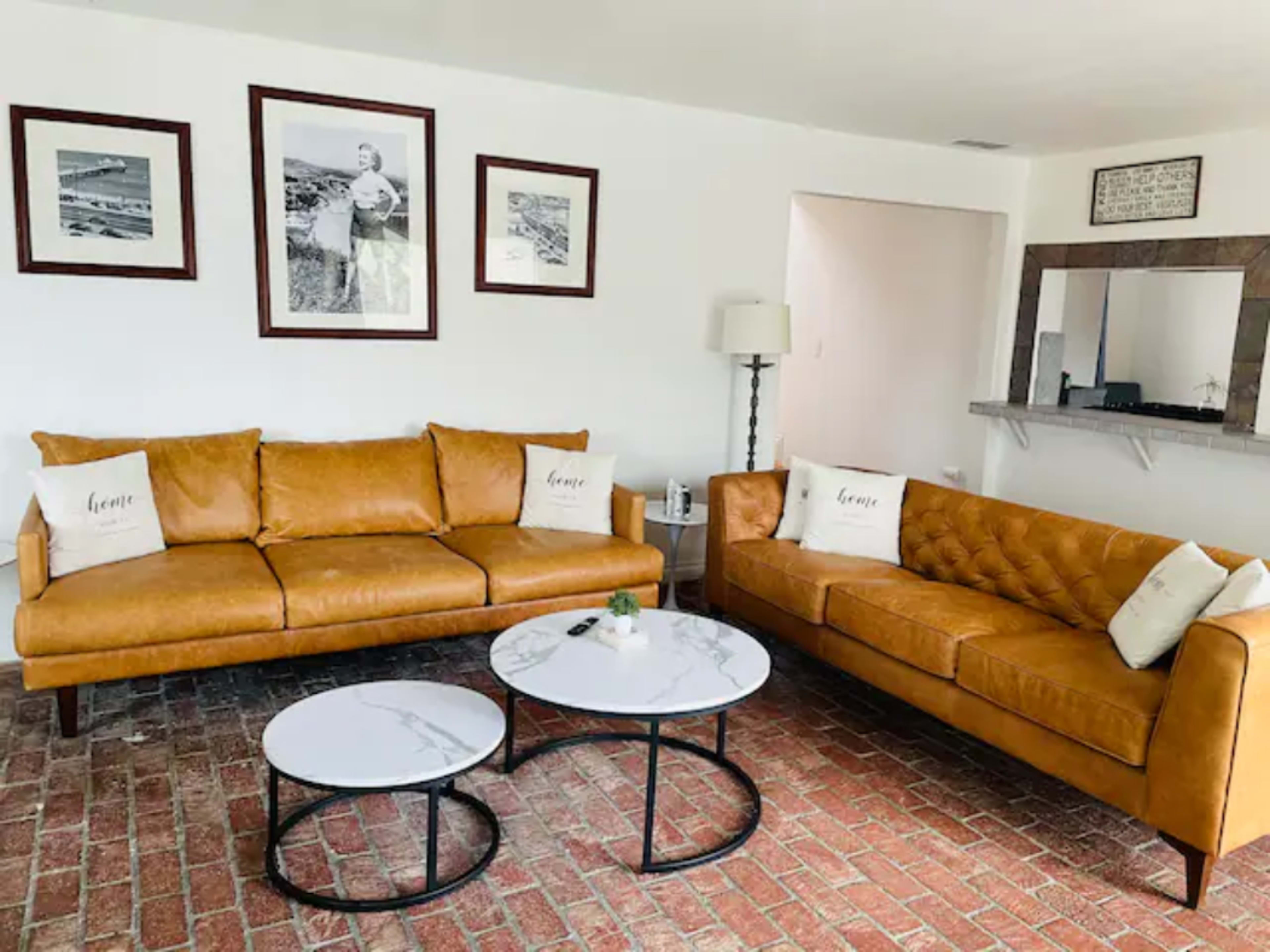 A living room with two leather sofas, framed black-and-white photographs on the wall, and two round marble-top coffee tables on a brick floor.
