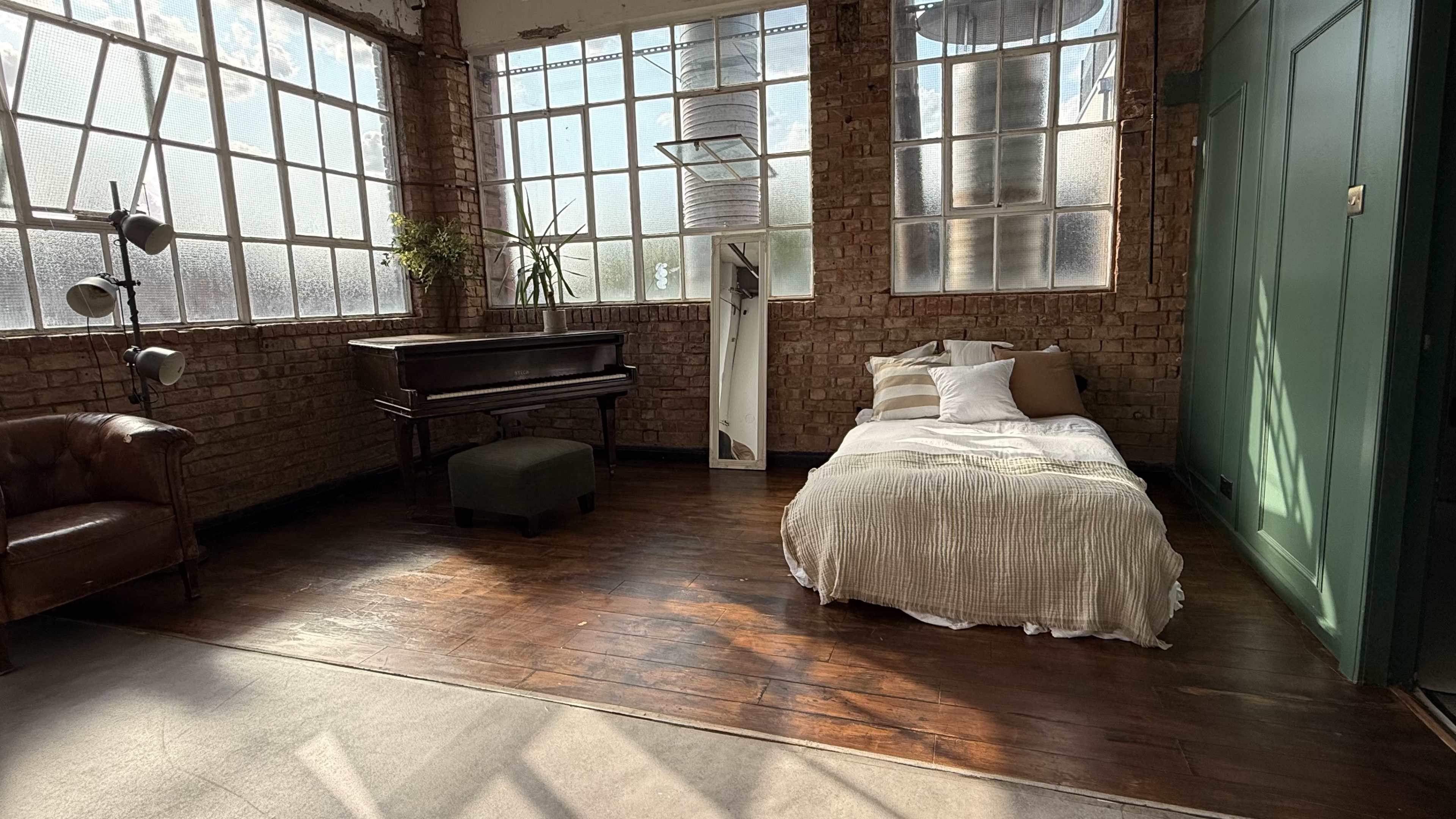 The image shows a spacious, well-lit room with large windows, a piano, a bed with a light-colored blanket, and a brown leather couch.