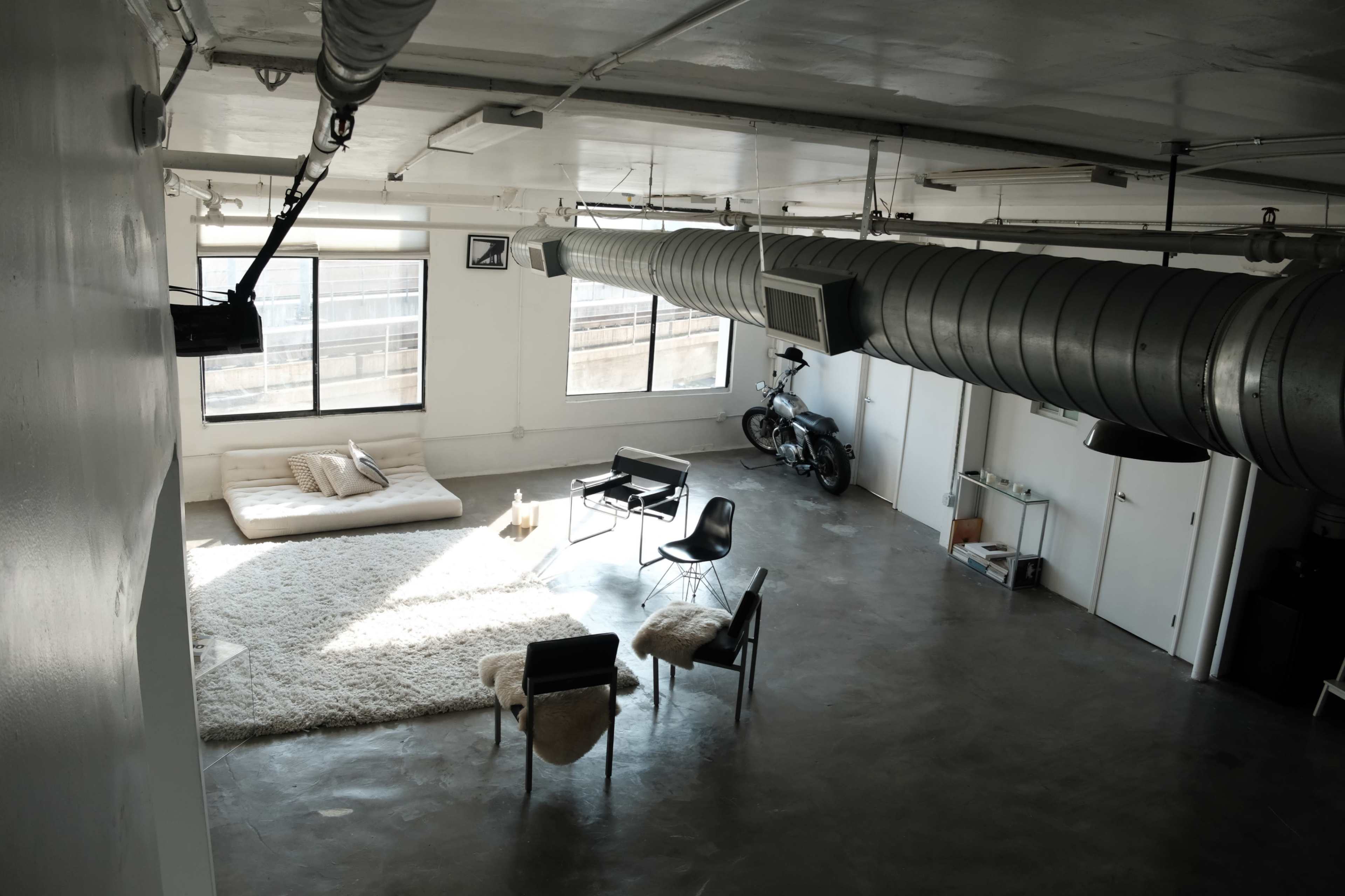 The image shows a spacious, minimalist loft interior with large windows, a white area rug, and a few pieces of furniture including a sofa, chairs, and a motorcycle parked against the wall.