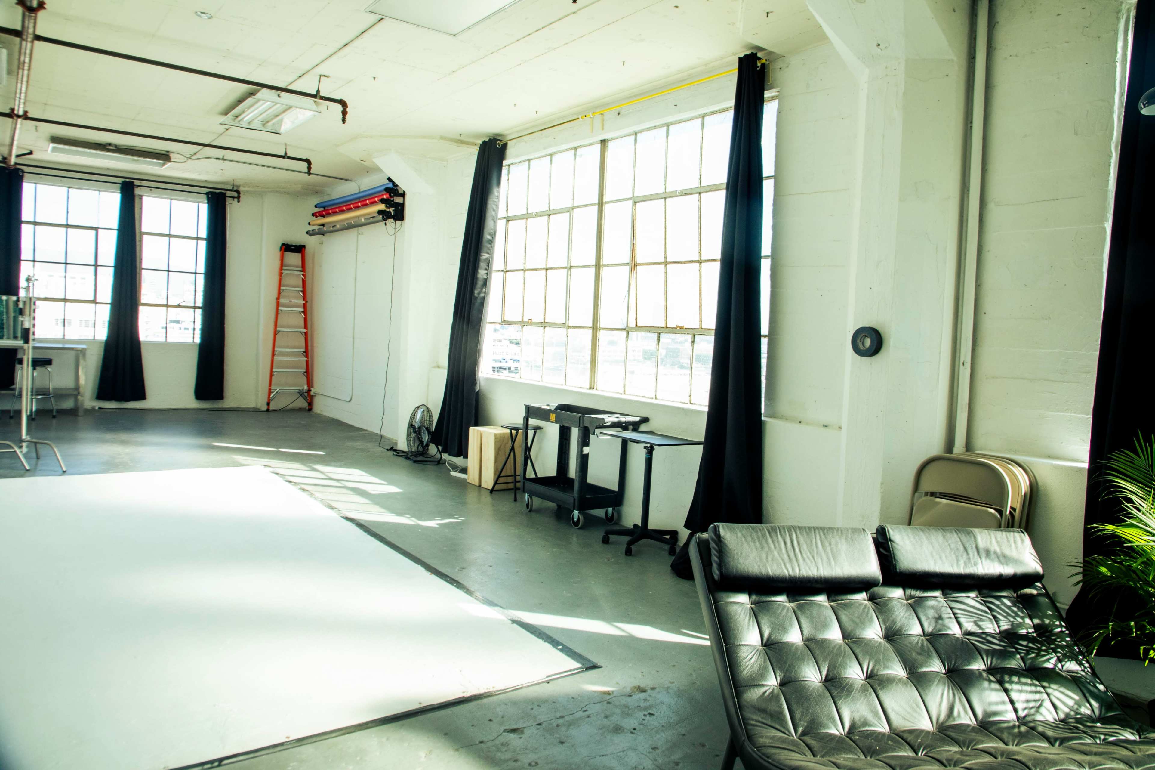 The image shows a bright, spacious studio with large windows, a black chaise lounge, a desk, and a stepladder in the background.