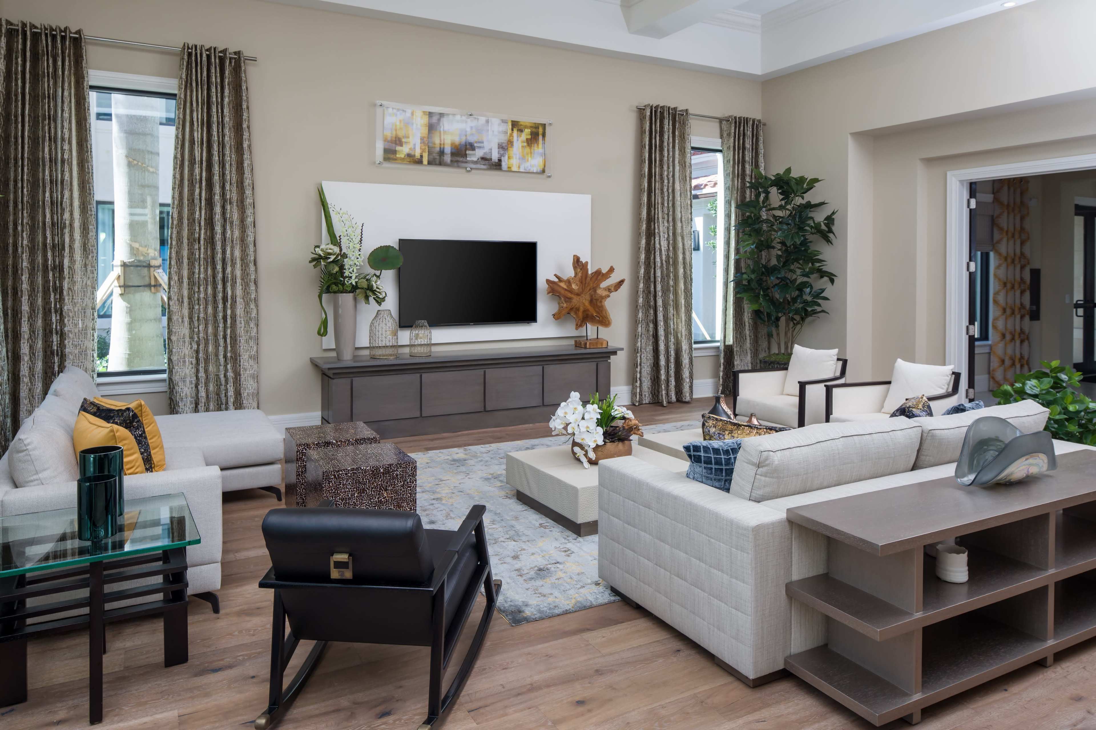 The image shows a modern living room featuring a large television mounted on a white wall, surrounded by various seating arrangements, decorative plants, and a central coffee table.