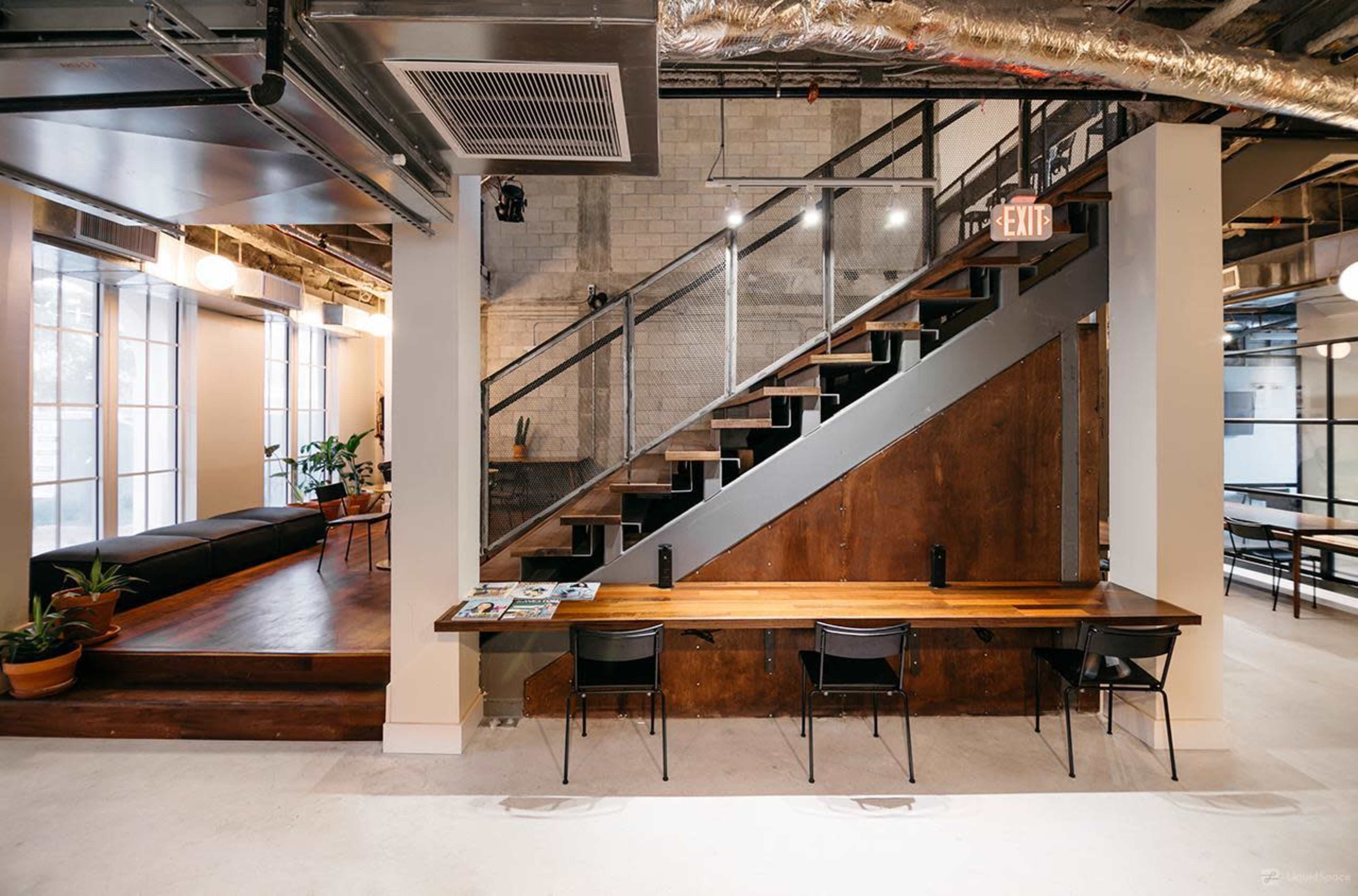 A contemporary office space with a wooden staircase, a lounge area with seating, and a long wooden table surrounded by chairs.