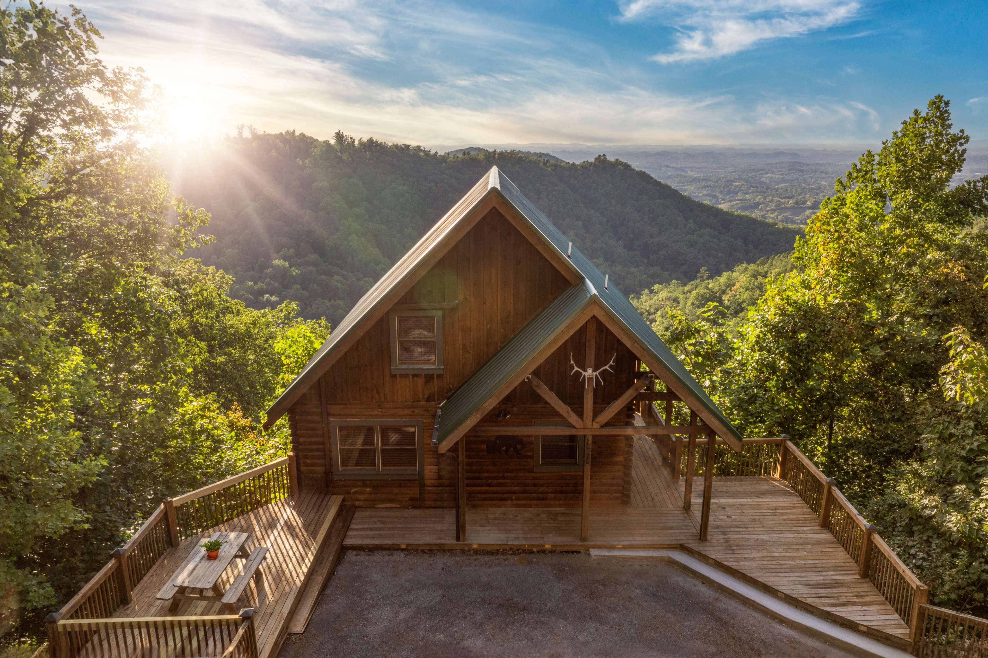 A wooden cabin with a green roof sits on a deck overlooking a valley and mountains, illuminated by a setting sun.