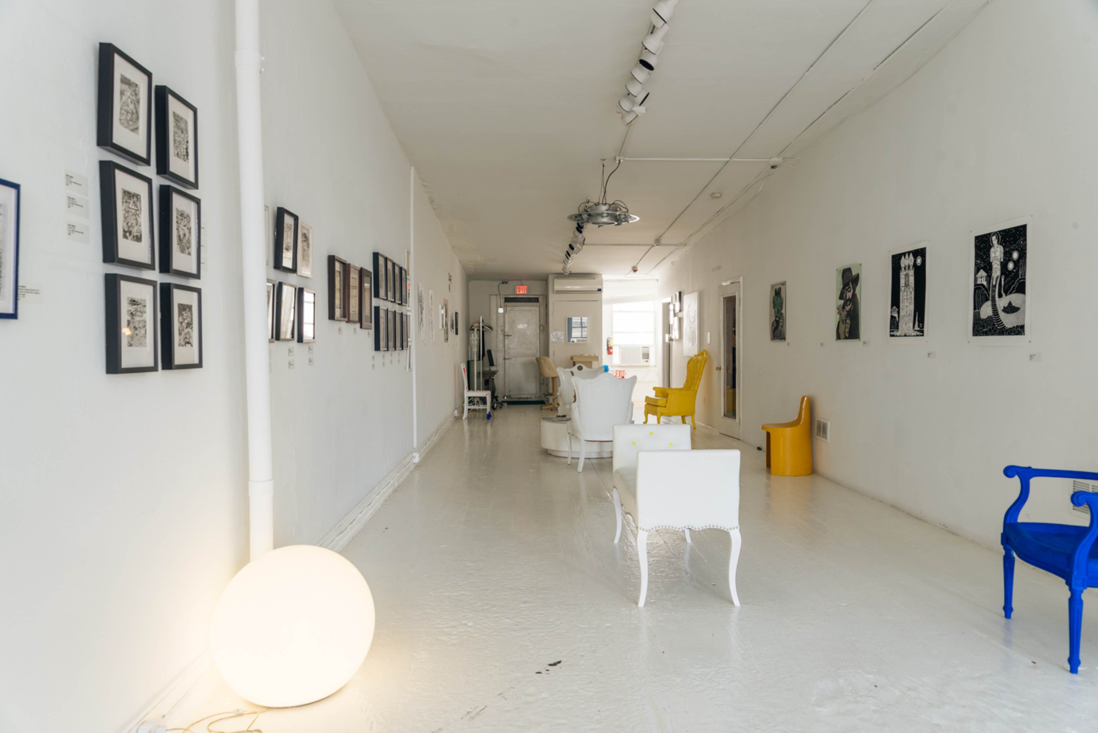 A long, white-walled gallery space featuring a series of framed artworks hung on the walls and various chairs positioned throughout the room.