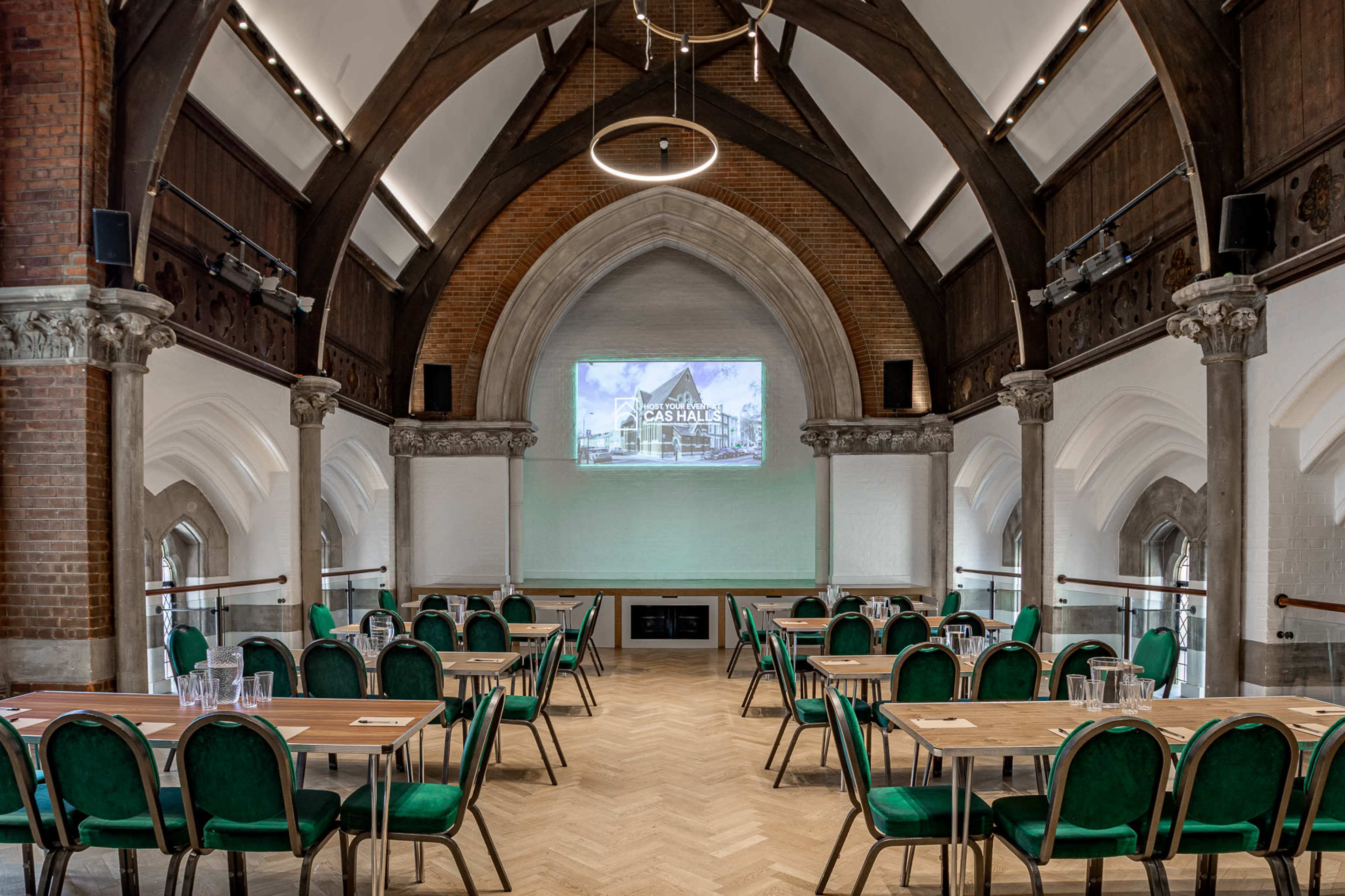 The image shows a spacious, architecturally designed room set up for a meeting, featuring rows of tables and green chairs, with a screen displaying a presentation at one end.