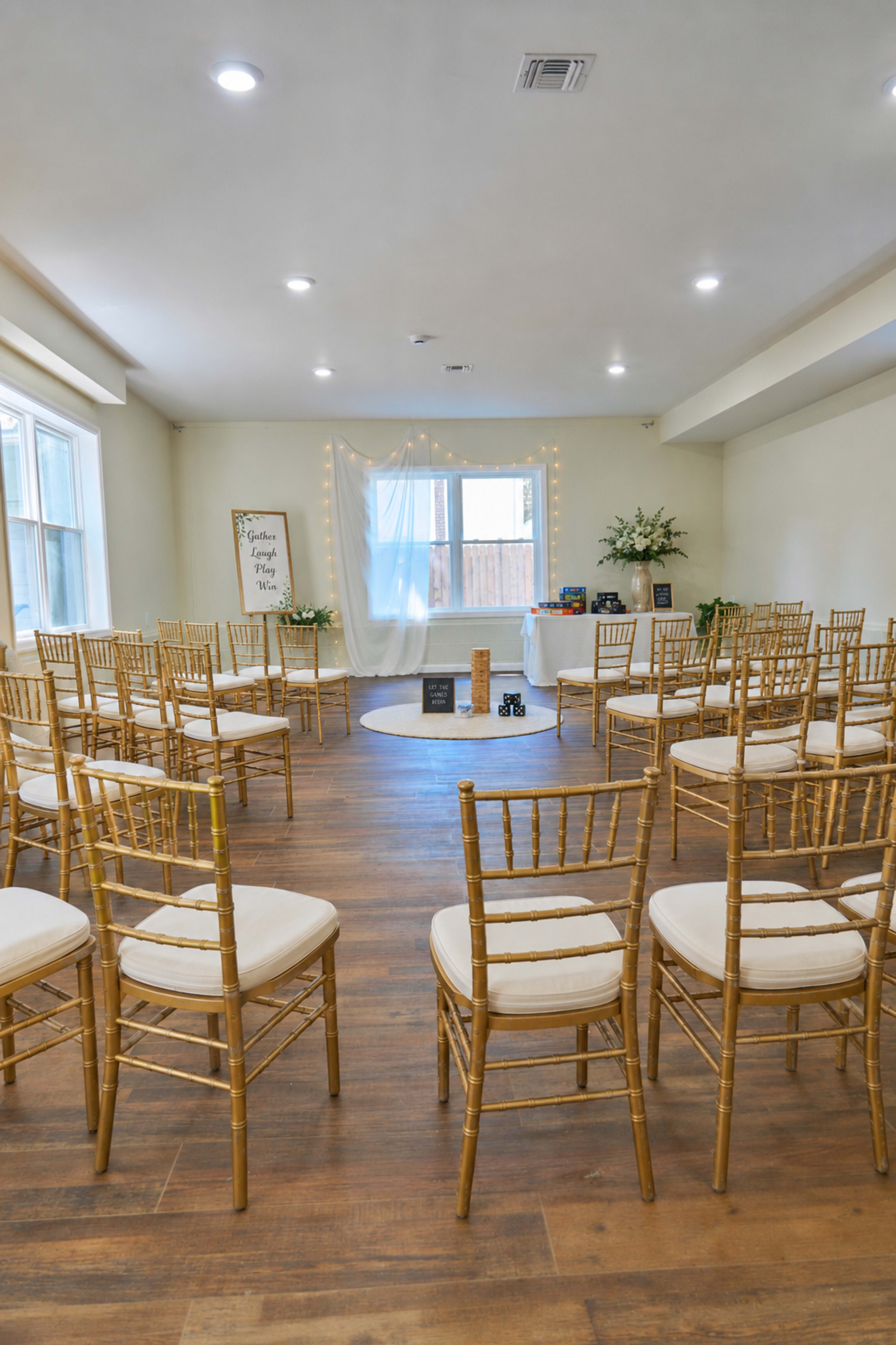 A small, elegantly arranged indoor space with rows of gold chiavari chairs facing a decorated ceremony area featuring a white backdrop and floral arrangements.