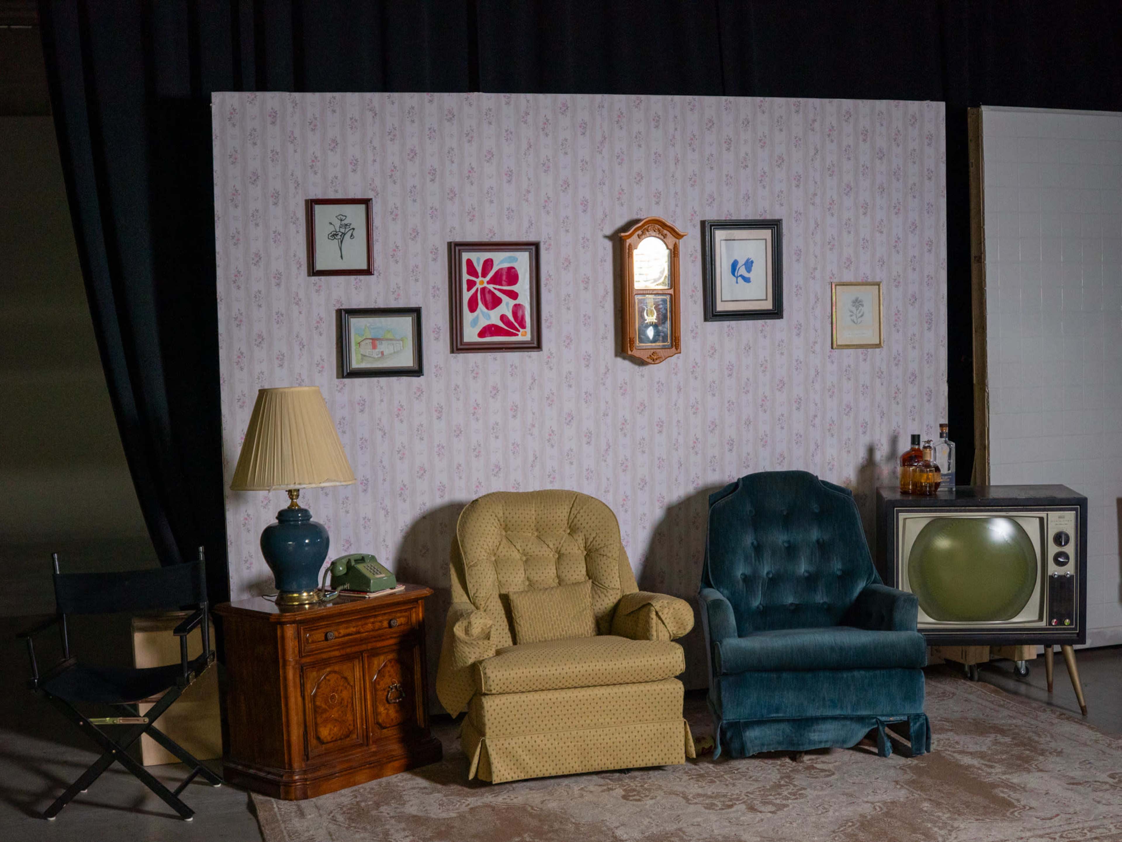 The scene features a vintage living room setup with two armchairs, a wooden side table, a lamp, a retro television, and framed artwork on the wall.