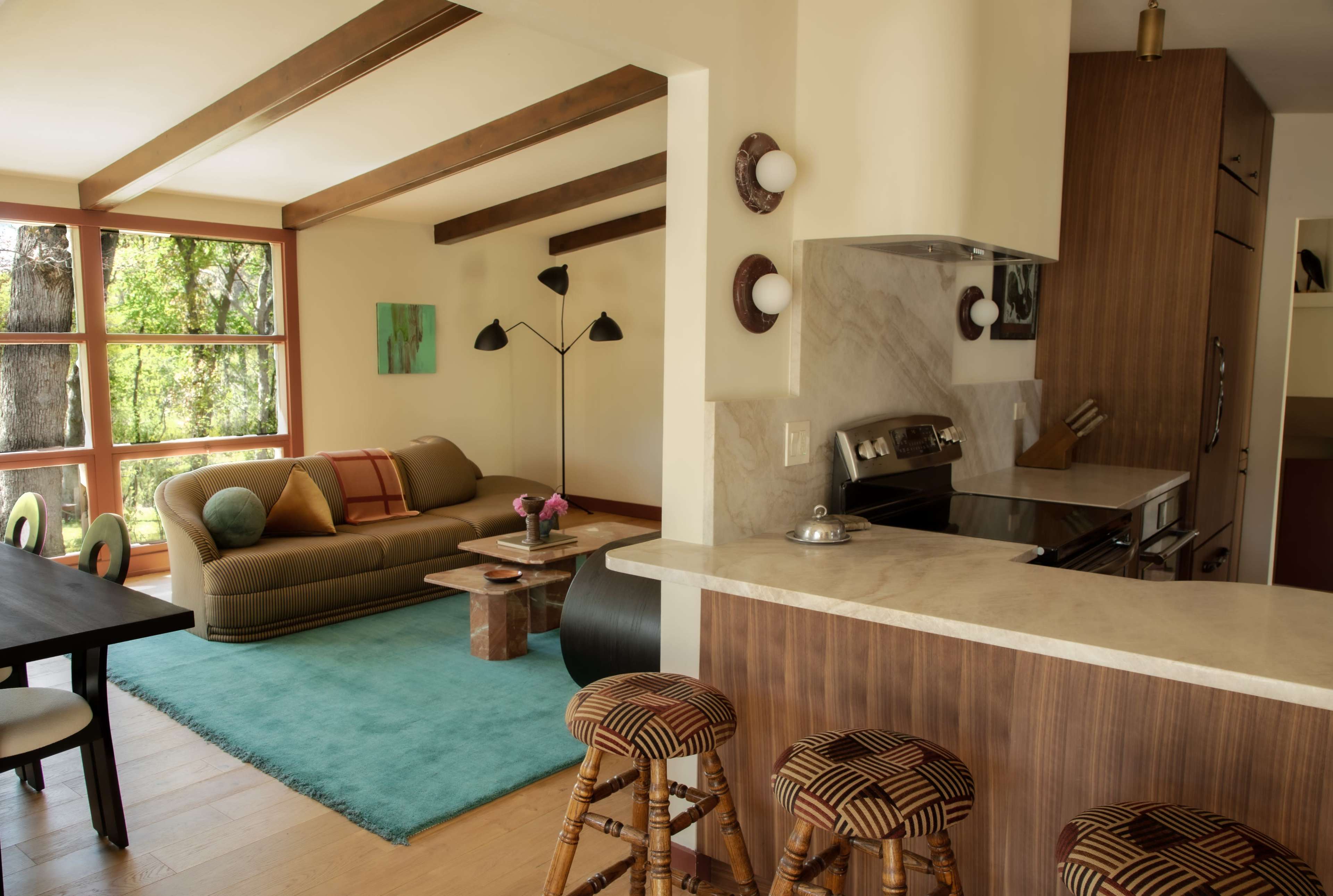 The image shows a modern living area with a seating arrangement, a dining table, and a kitchen bar, all set in a bright space that features wooden beams and large windows.