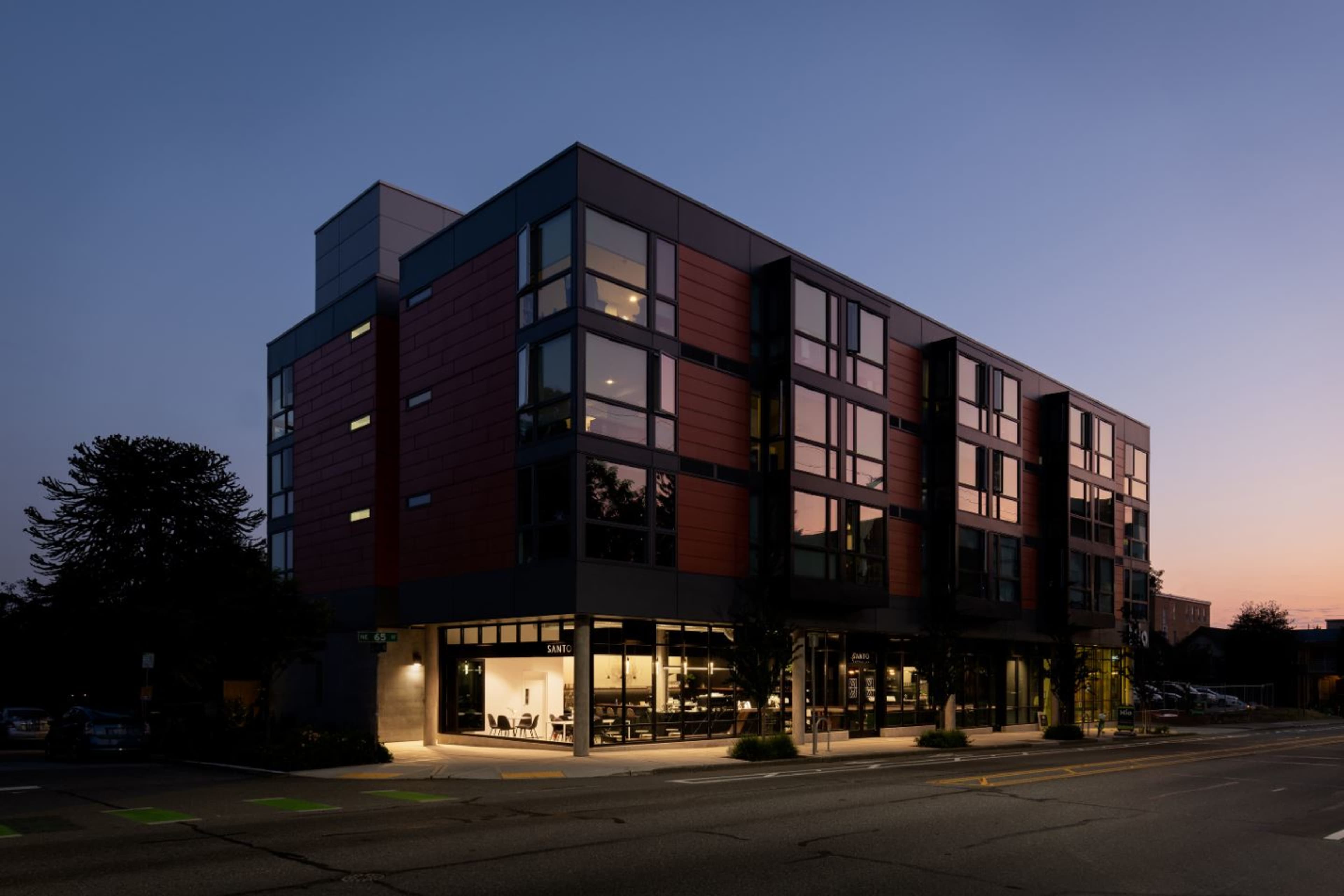 A modern multi-story building with large windows and a mix of red and gray exterior finishes stands illuminated at dusk.