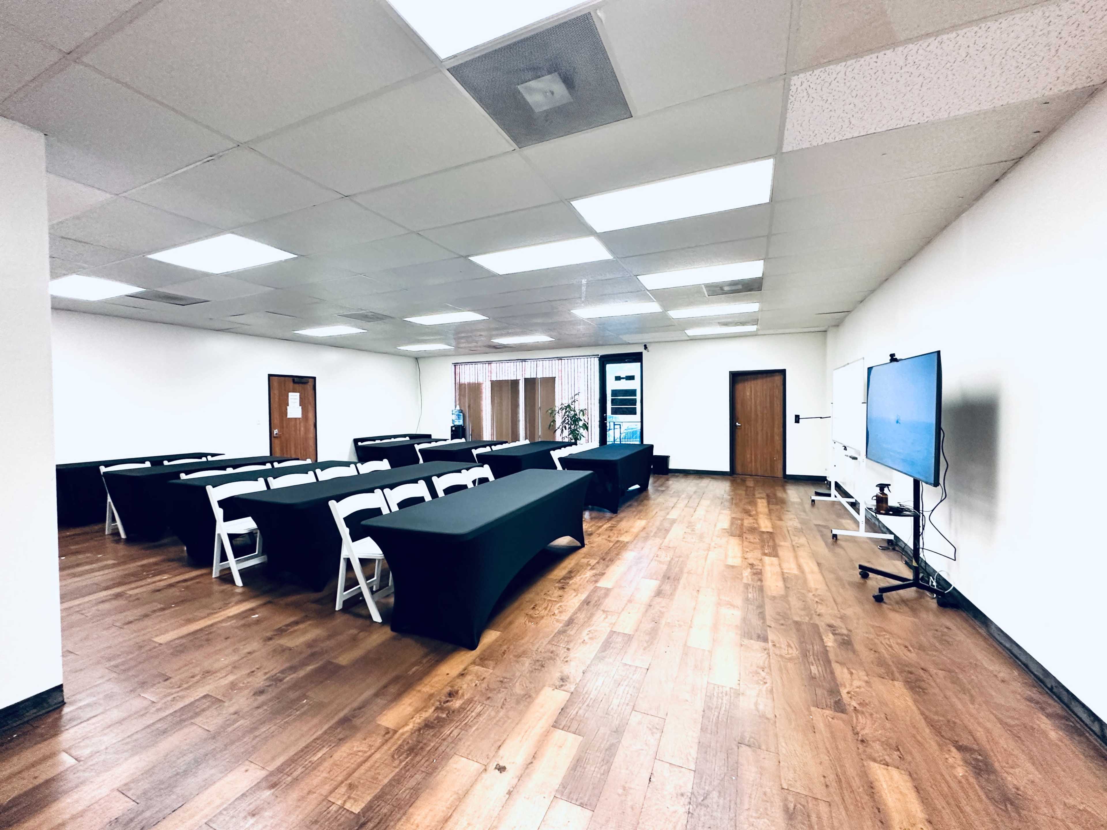 A large meeting room with rows of black-clothed tables, white chairs, and a mounted television on the wall.