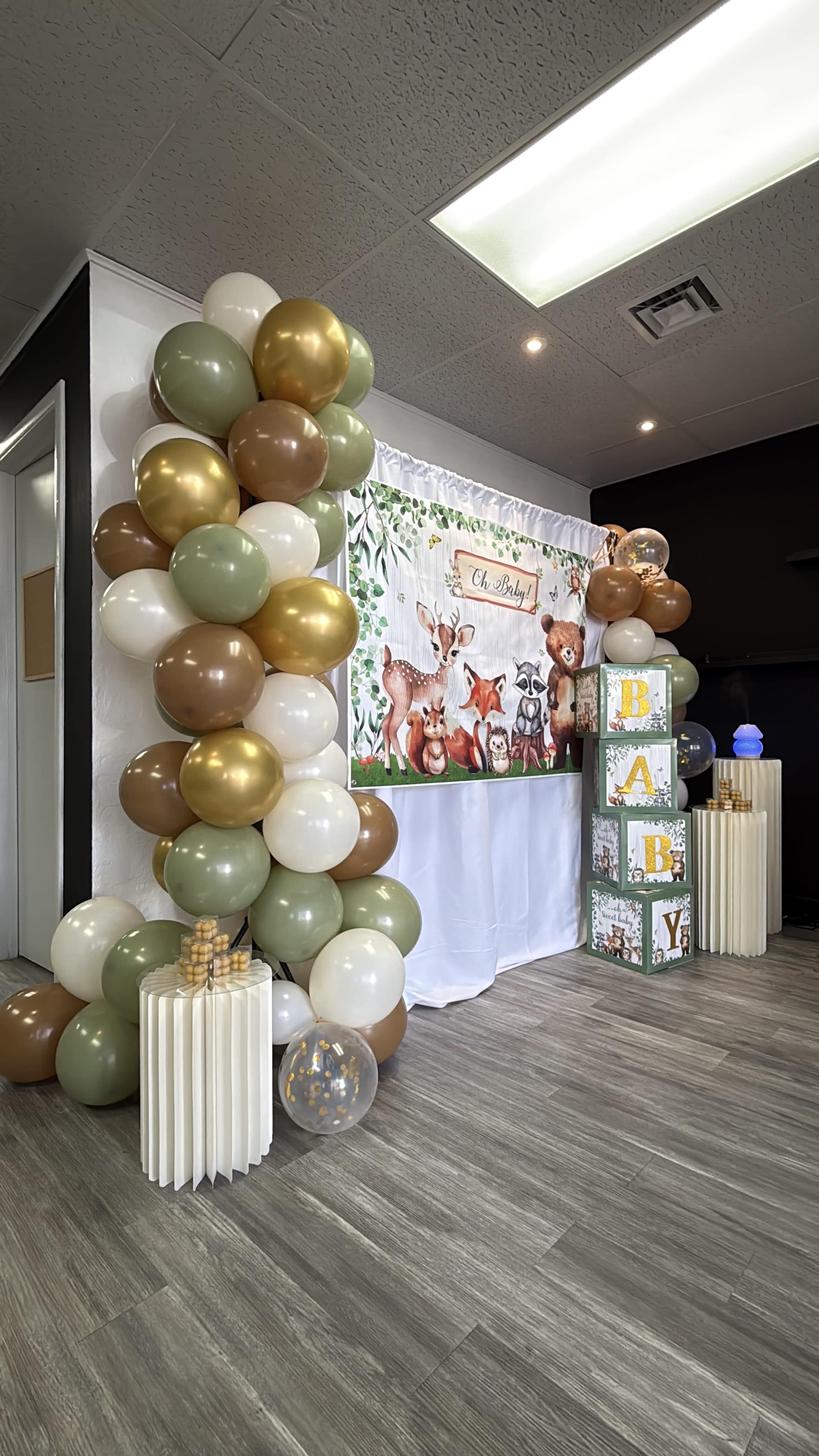 The image shows a decorated area with a backdrop featuring woodland animals, colorful balloons in shades of brown, gold, and green, and large blocks spelling out "BABY."