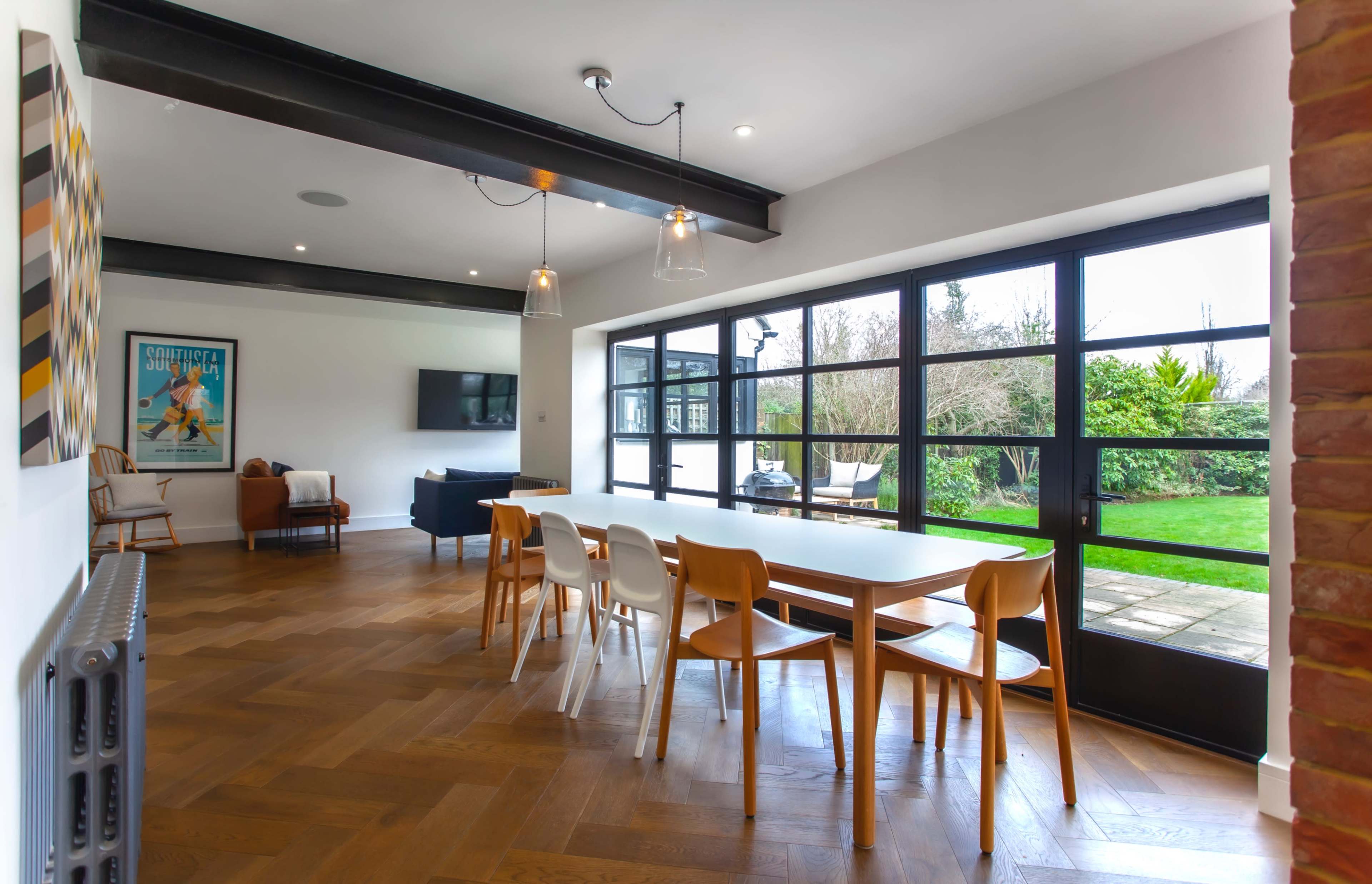 The image shows a modern dining area with a large table and light-colored chairs, open to a living space featuring a TV and large windows overlooking a garden.