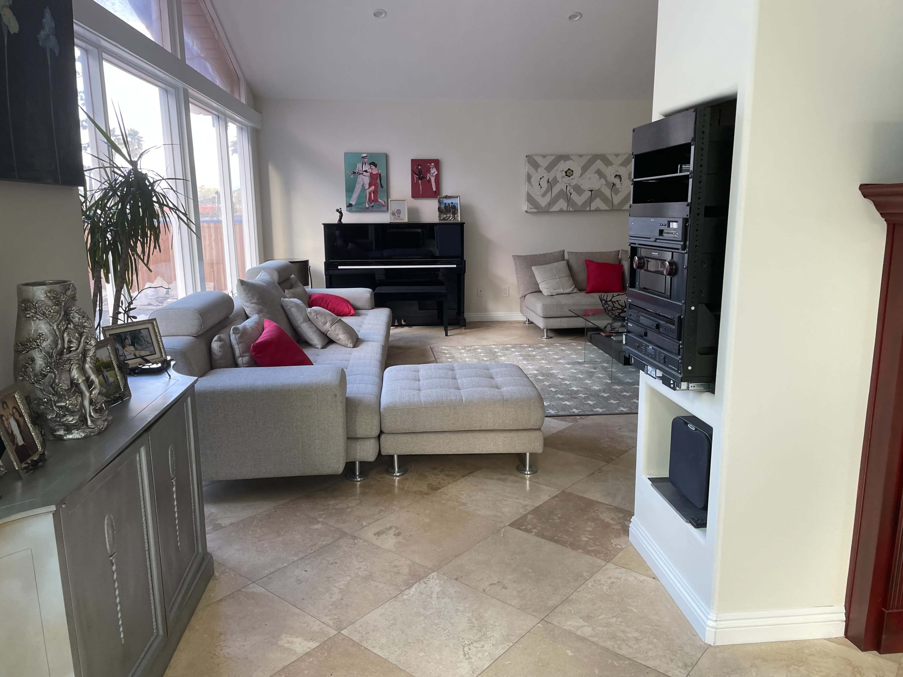 The image shows a bright living room with a sectional sofa, a piano, and a decorative wall featuring artwork.
