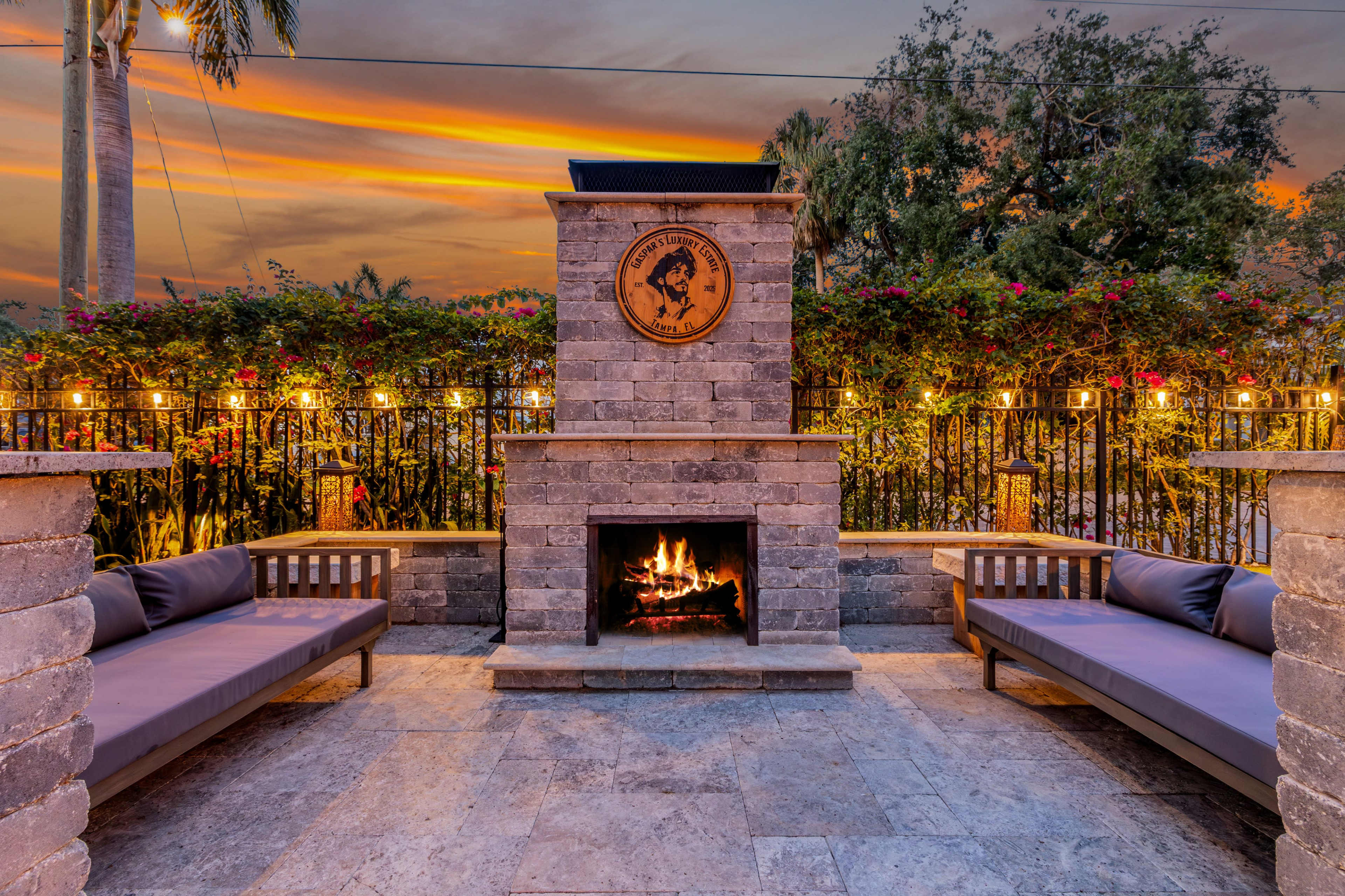 The image depicts a cozy outdoor seating area with two stone benches facing a fire pit, surrounded by lush greenery and string lights against a sunset sky.