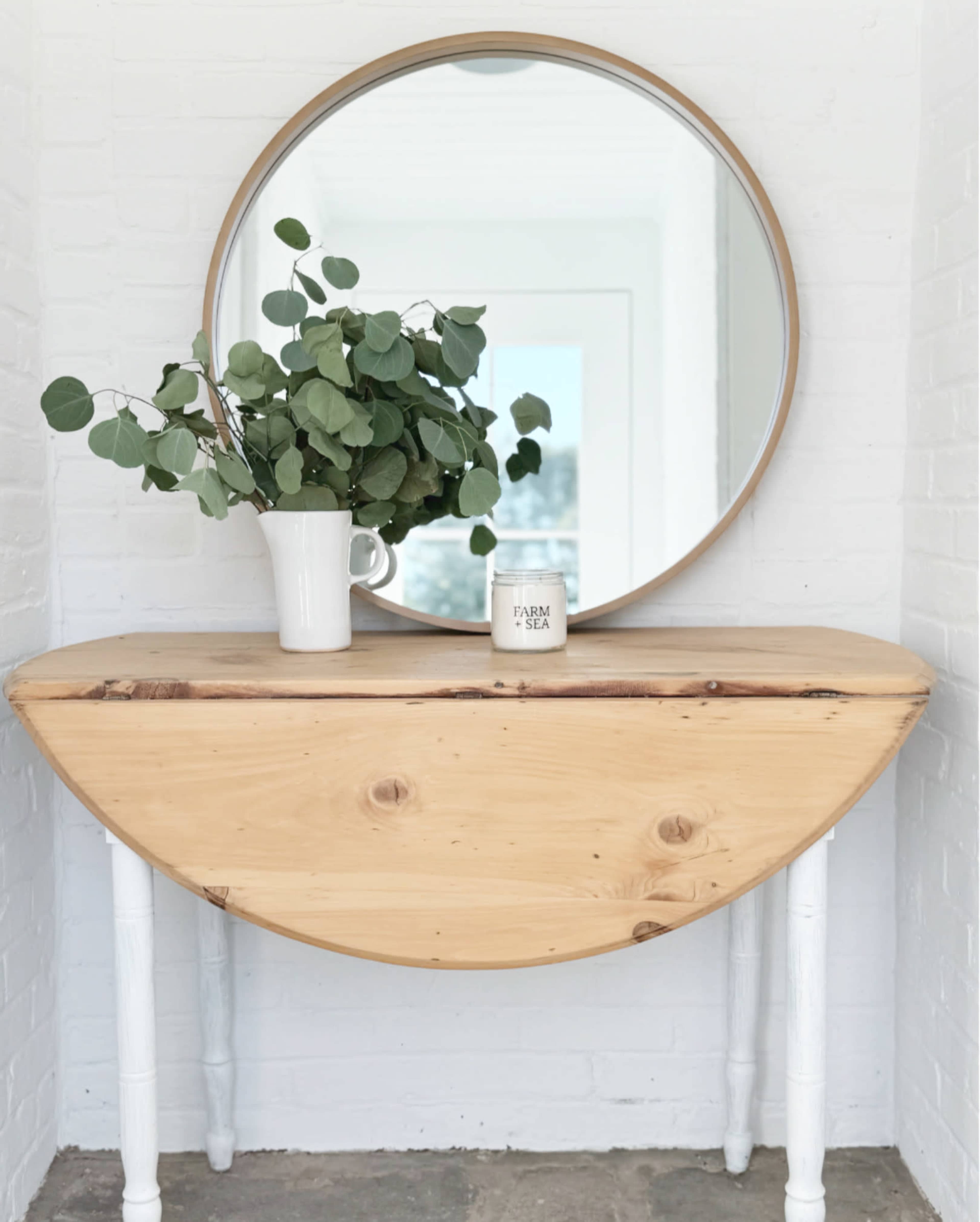 A wooden half-moon table with white legs holds a vase of eucalyptus and a candle, positioned beneath a round mirror against a white brick wall.