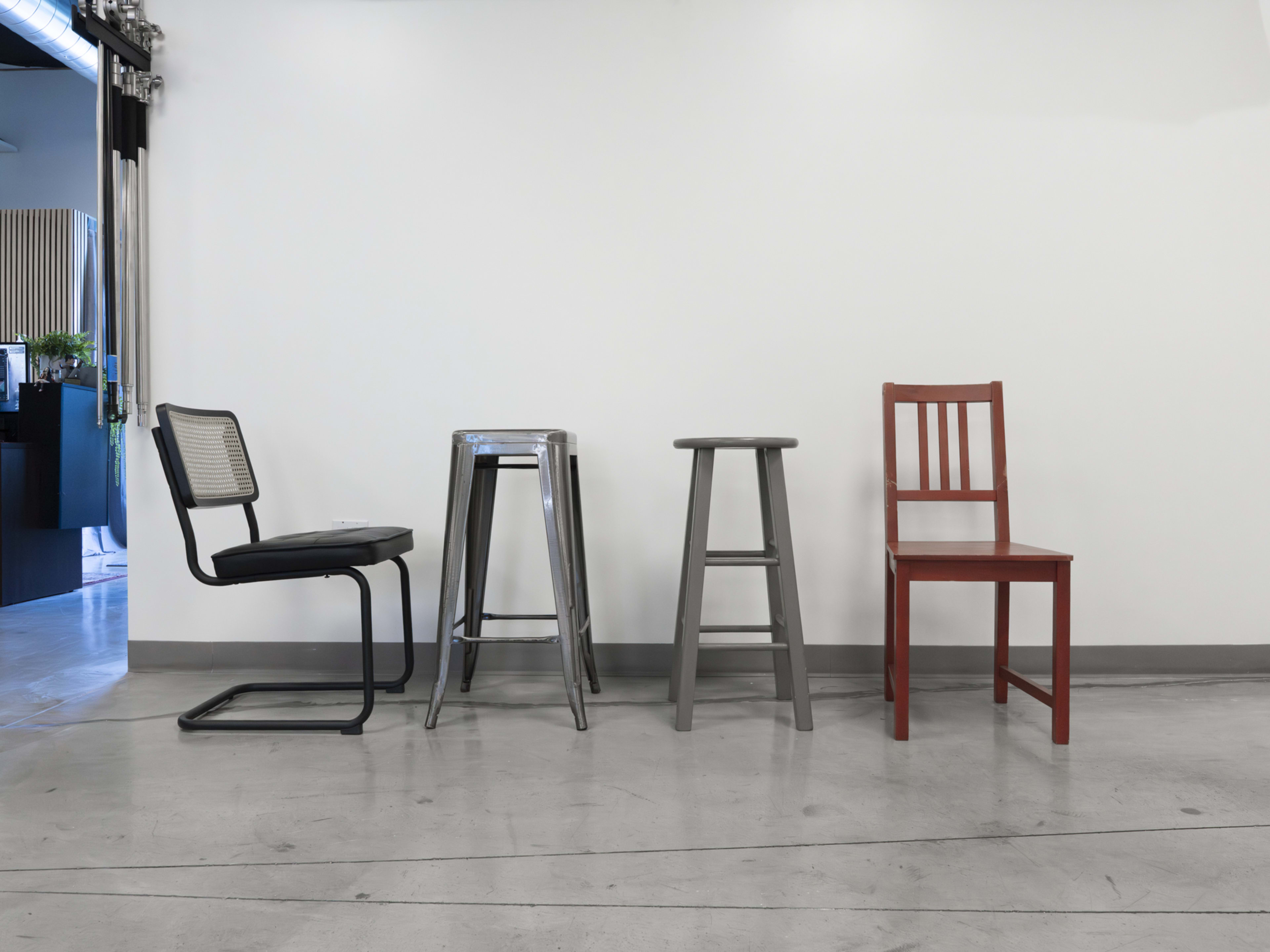 The image shows four different types of chairs arranged in a row against a white wall in a minimalist setting.