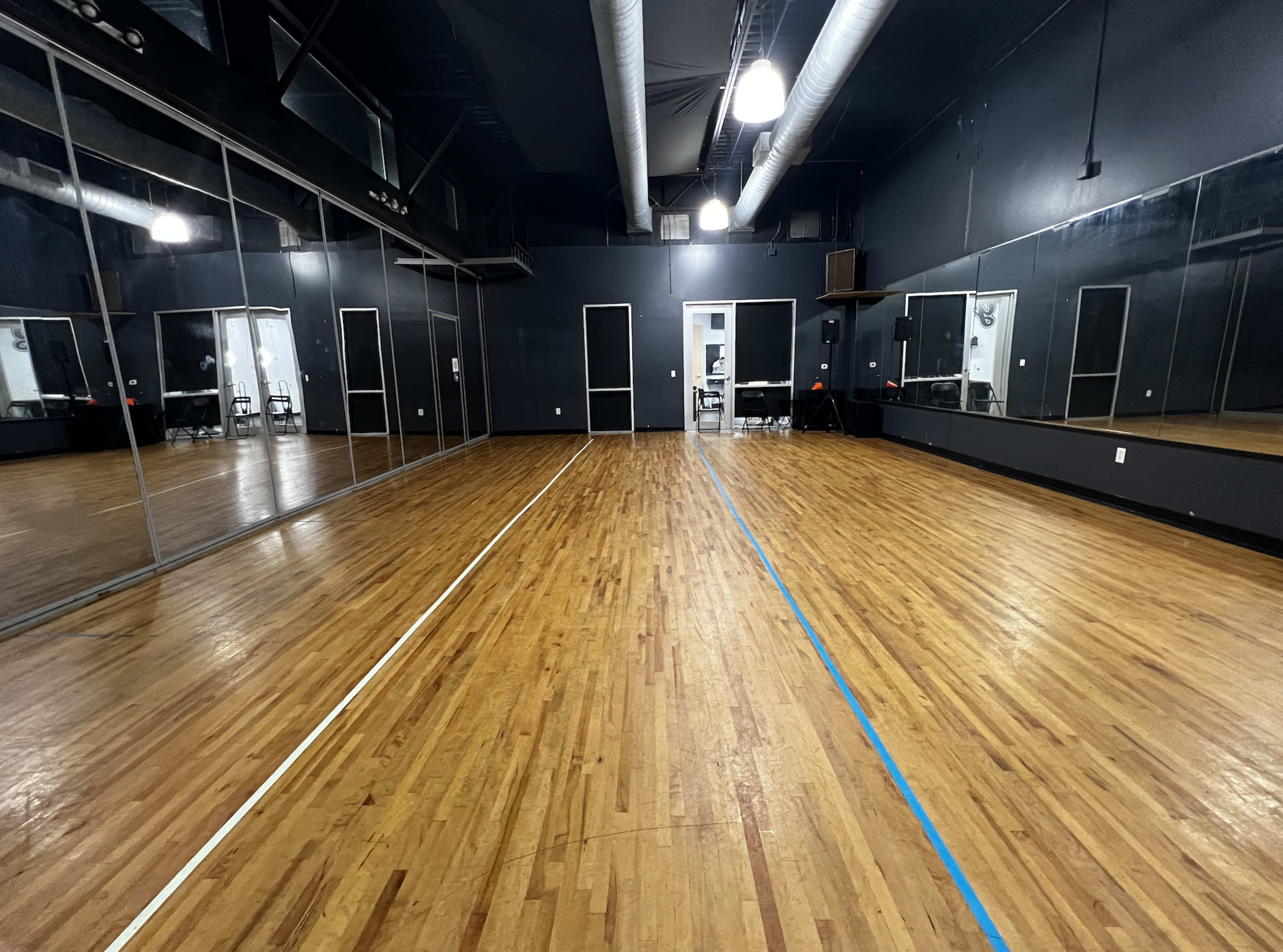 An empty dance studio with wooden floors, large mirrors on one wall, and blue and white lines marking the floor.