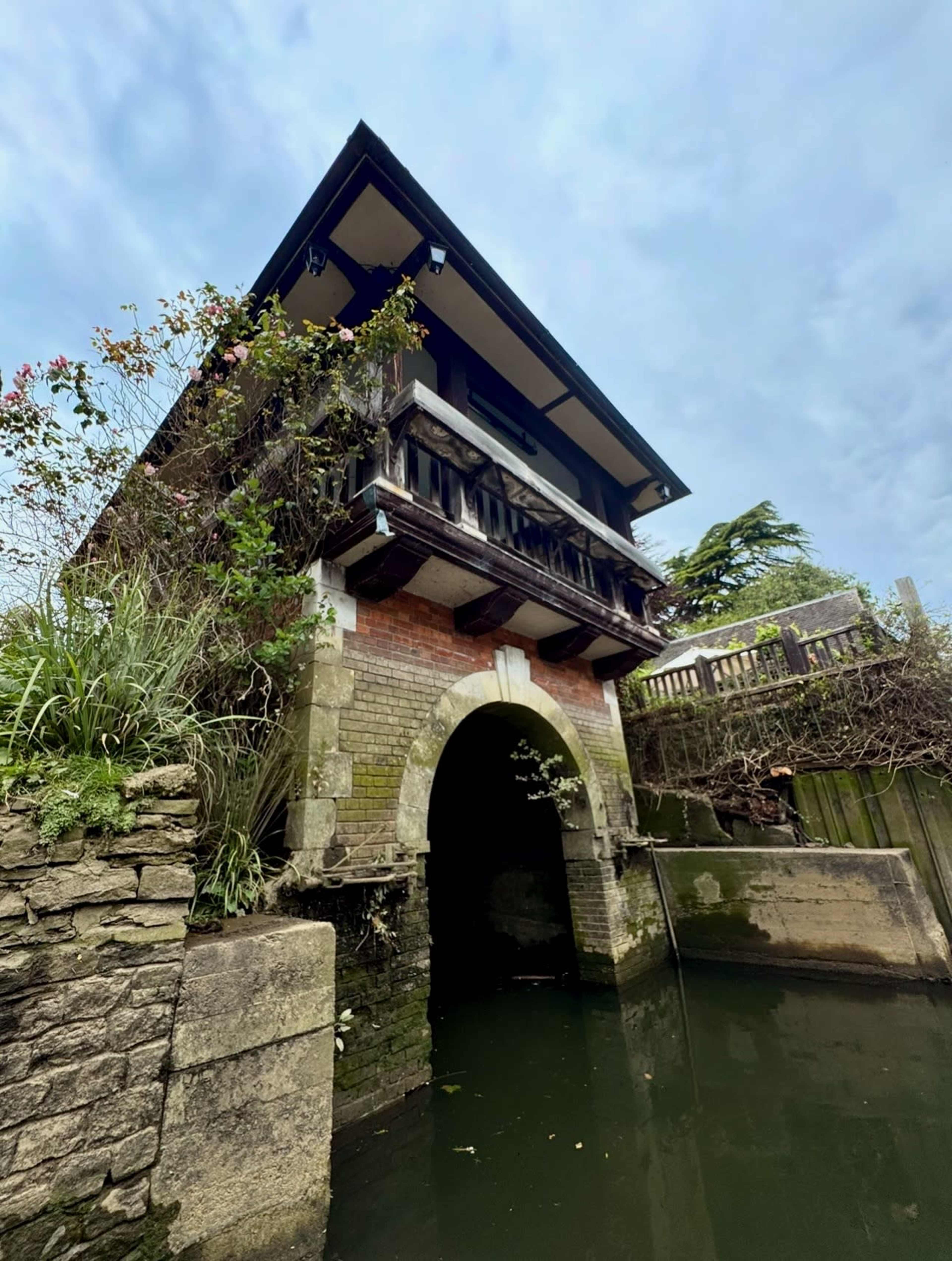 A building with a cantilevered balcony stands above a waterway, with stone walls and overgrown greenery surrounding it.