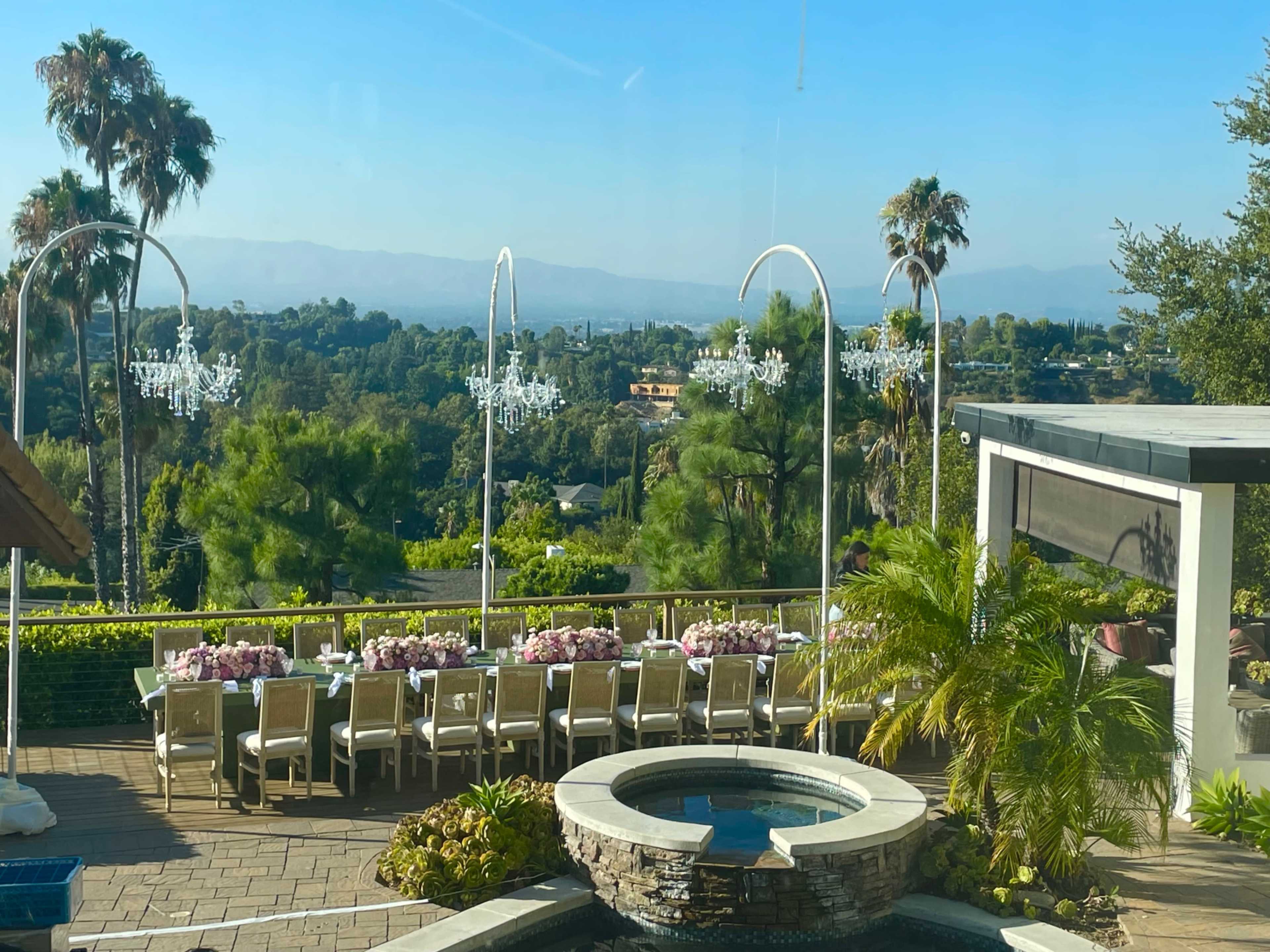 A landscaped terrace features a long dining table with chairs, overlooking a distant valley and mountains, with palm trees and decorative chandeliers in the background.