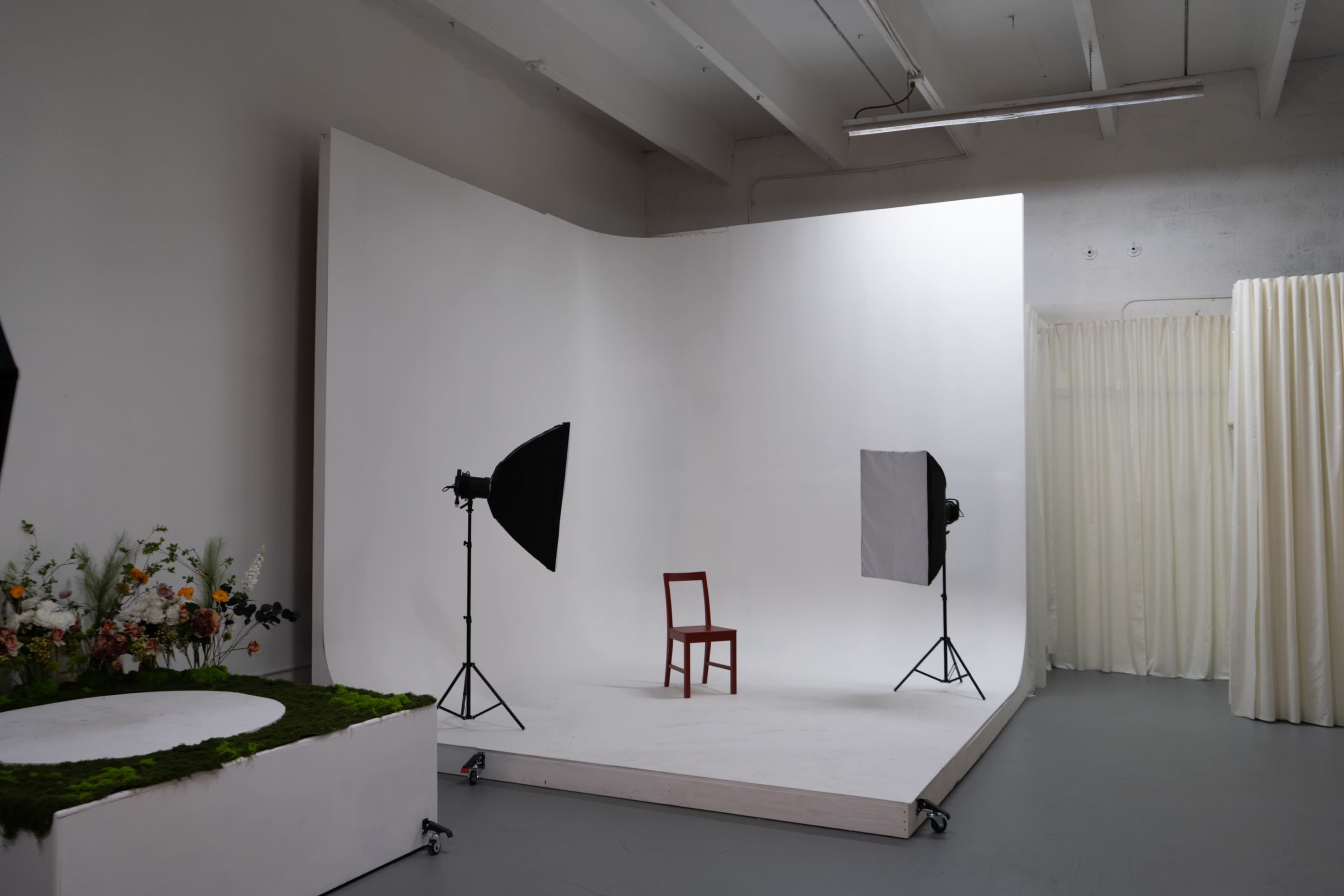 The image shows a minimalist photography studio with a single wooden chair positioned on a white platform, flanked by two softbox lights and a floral arrangement on one side.